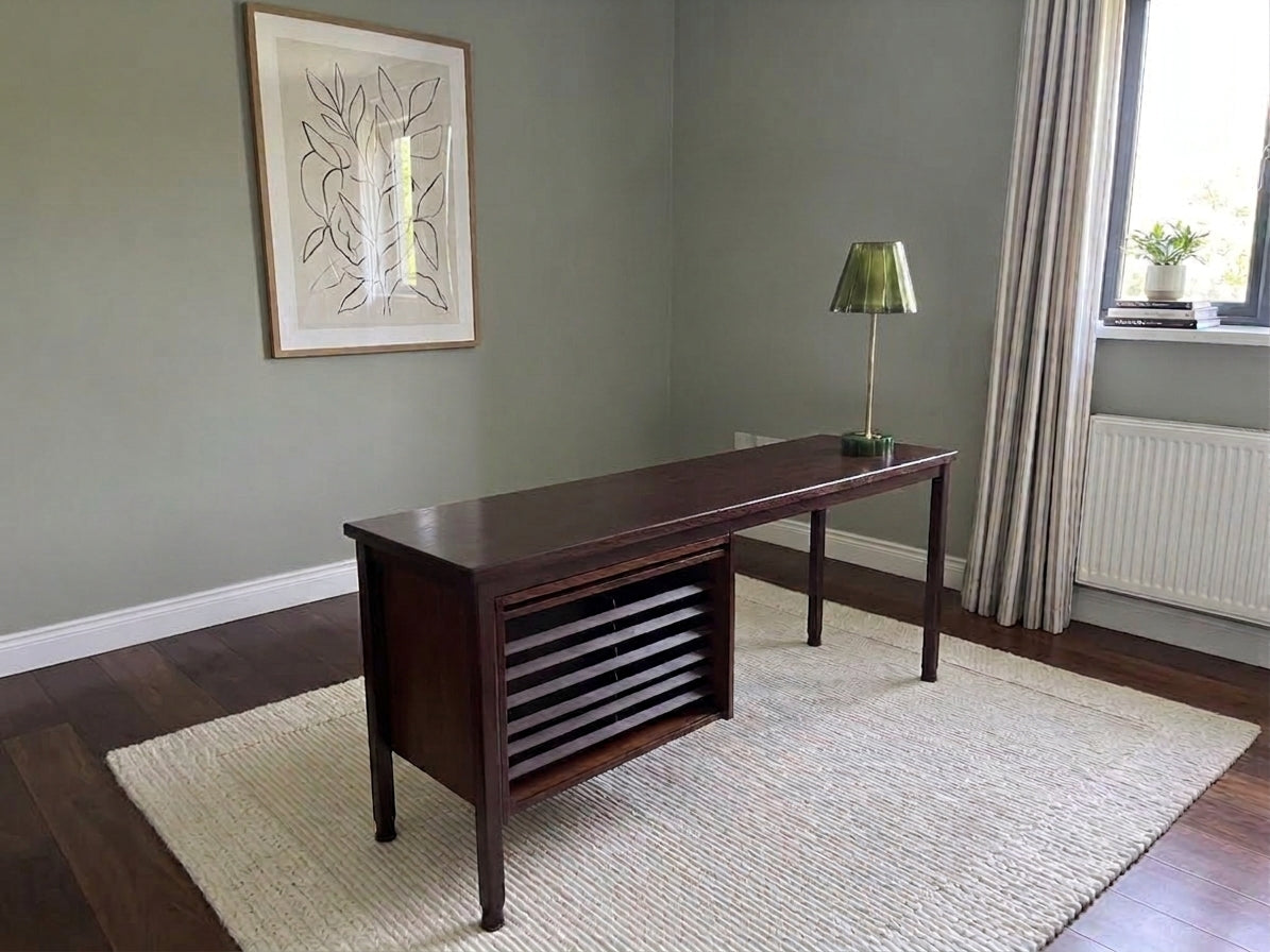 A dark brown wooden writing desk with a slatted lower cabinet, placed on a cream woven rug in a room with sage green walls, a window with curtains, and a green lamp on top.