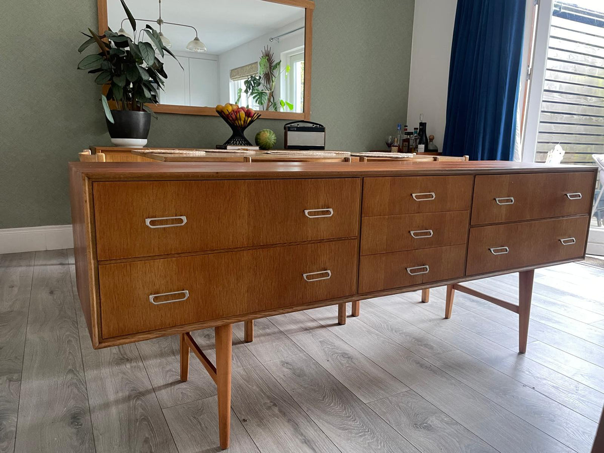 Mid-century modern wooden meredew credenza with six drawers and silver handles, shown in a styled room with gray wood floors, a green wall, and a mirror behind it.