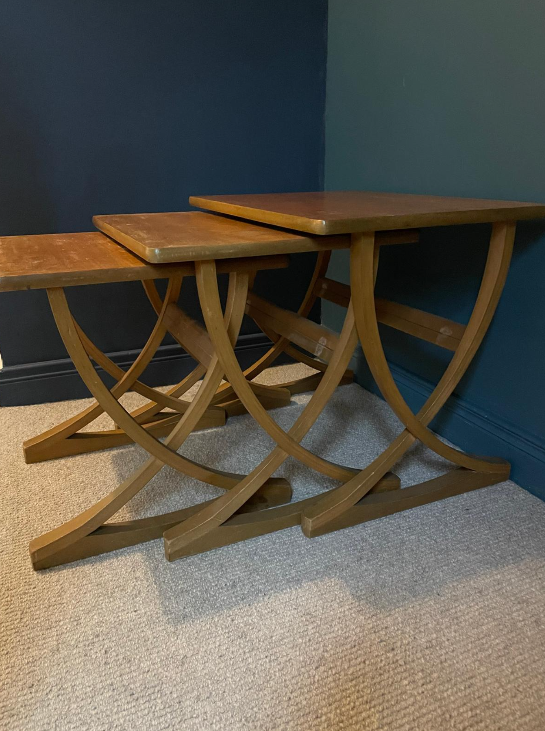 Three nested wooden nesting tables with a warm medium-brown finish, set on a beige carpet against a dark teal wall.