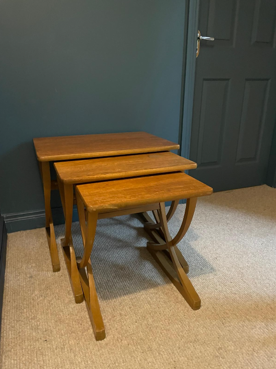 Three nested wooden nesting tables with a warm medium-brown finish, set on a beige carpet against a dark teal wall.