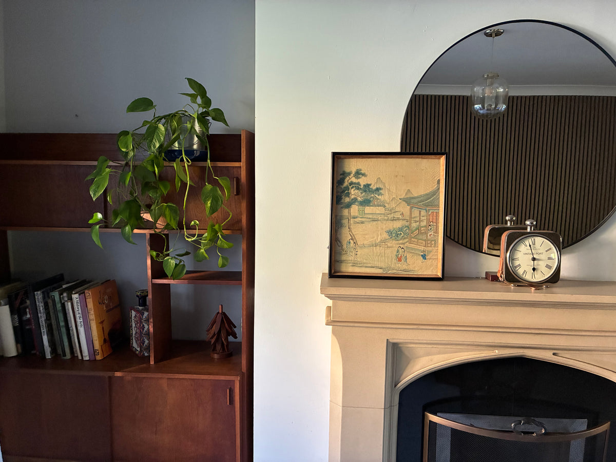 A framed vintage-style landscape print with an Asian-inspired pagoda scene, displayed on a cream-colored fireplace mantel next to a retro alarm clock and beneath a large round mirror. To the left, a wooden bookshelf holds books and a green trailing plant.
