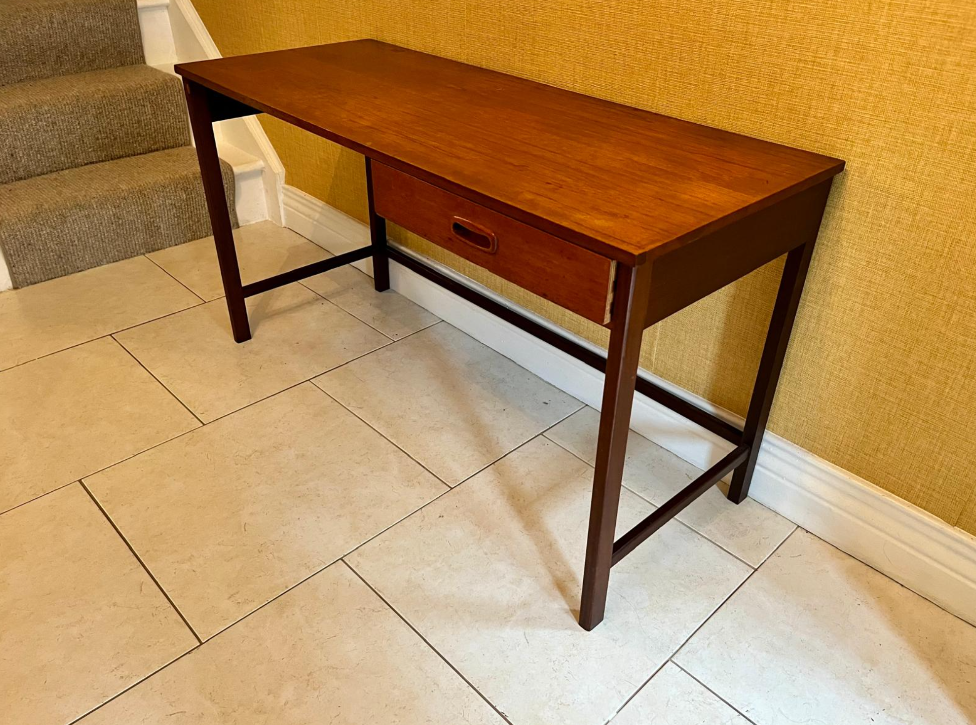 A mid-century modern wooden writing desk with a single drawer, positioned in a home setting against a textured yellow wall and tiled floor.