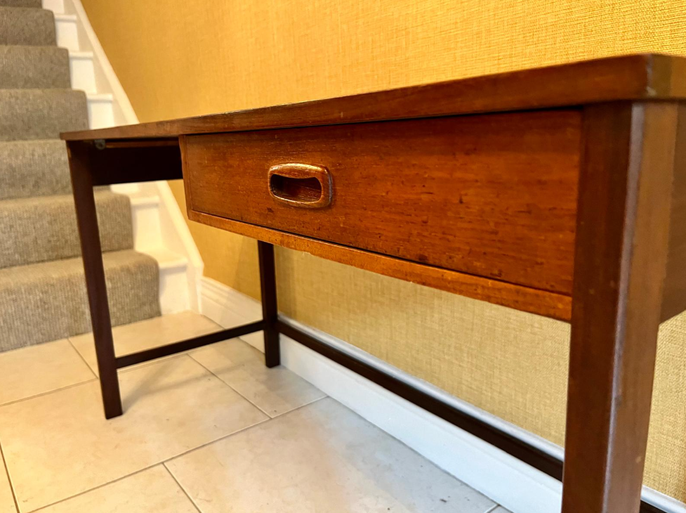 A mid-century modern wooden writing desk with a single drawer, positioned in a home setting against a textured yellow wall and tiled floor.