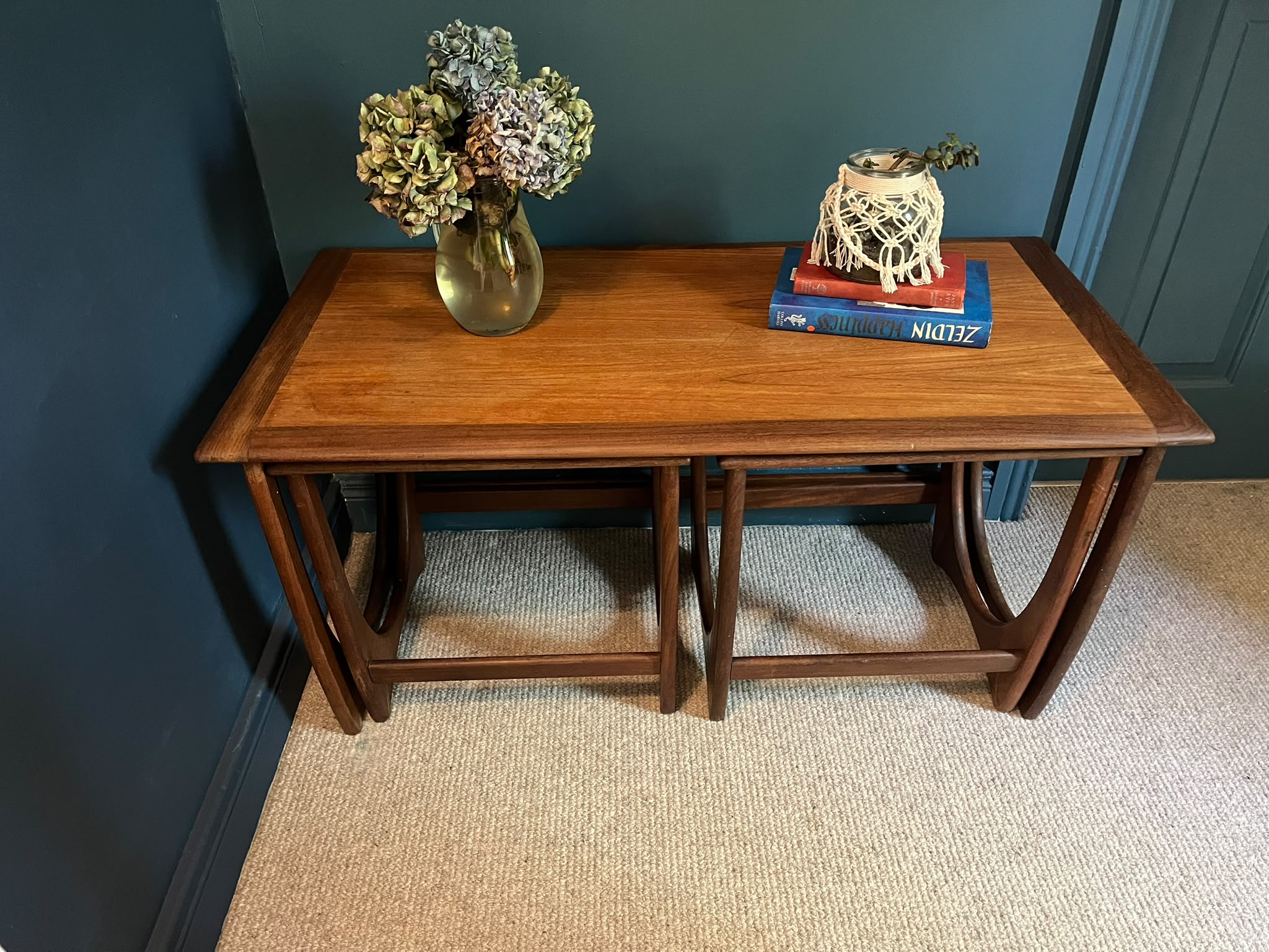 A G Plan Astro coffee nest set with a warm medium-brown finish, styled with a gold vase of dried flowers and a decorative sculpture on books, set against a dark teal wall and beige carpet by Grace & Joy Furniture