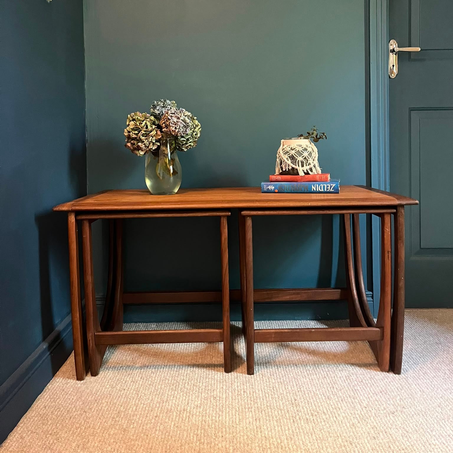 A G Plan Astro coffee nest set with a warm medium-brown finish, styled with a gold vase of dried flowers and a decorative sculpture on books, set against a dark teal wall and beige carpet by Grace & Joy Furniture