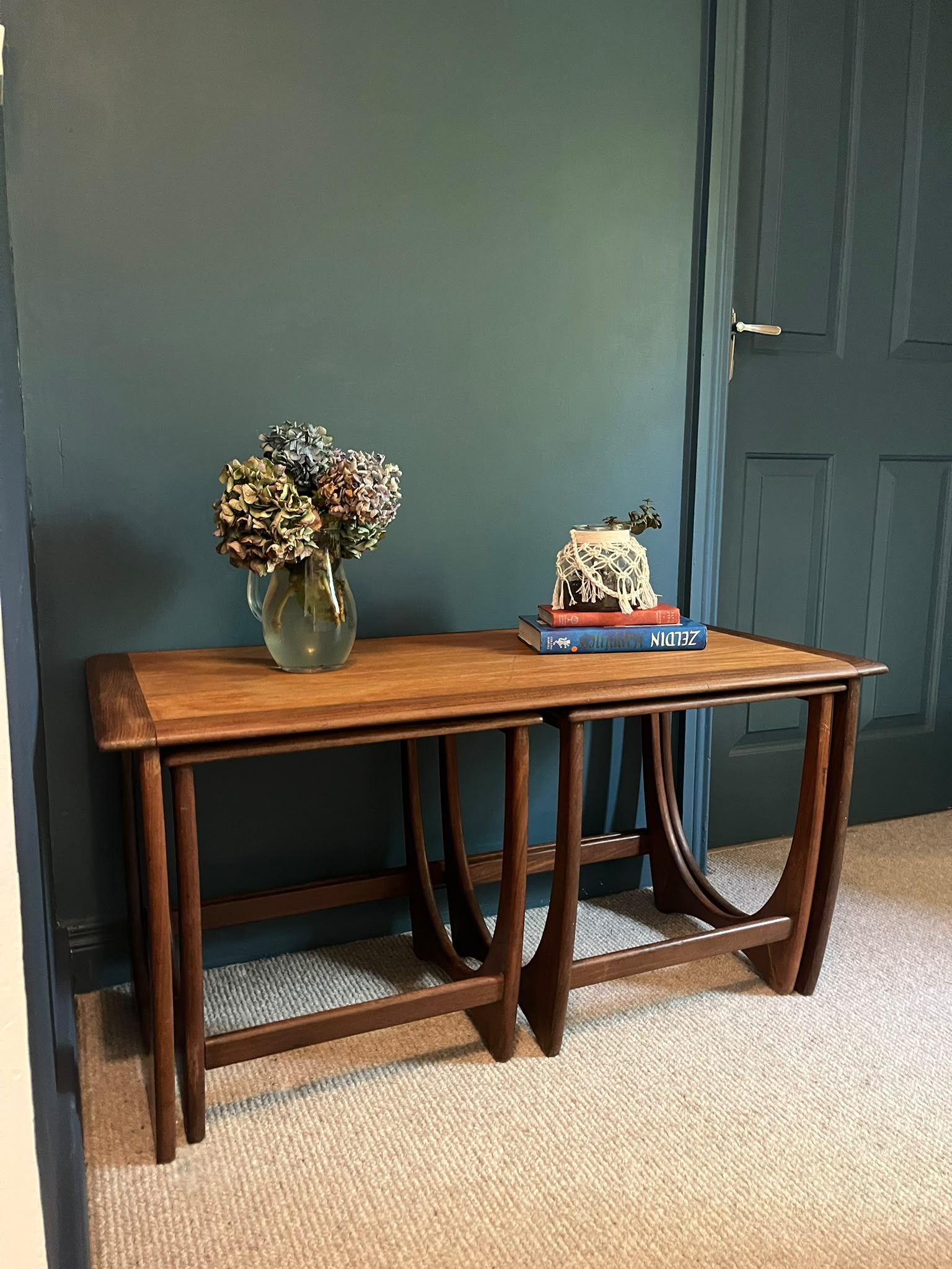 A G Plan Astro coffee nest set with a warm medium-brown finish, styled with a gold vase of dried flowers and a decorative sculpture on books, set against a dark teal wall and beige carpet by Grace & Joy Furniture