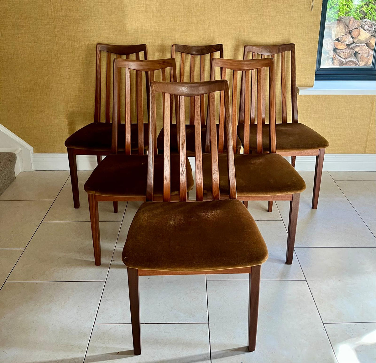 Six mid-century G Plan modern wooden dining chairs with vertical slat backs and brown velvet seats, arranged on a tiled floor against a textured yellow wall.