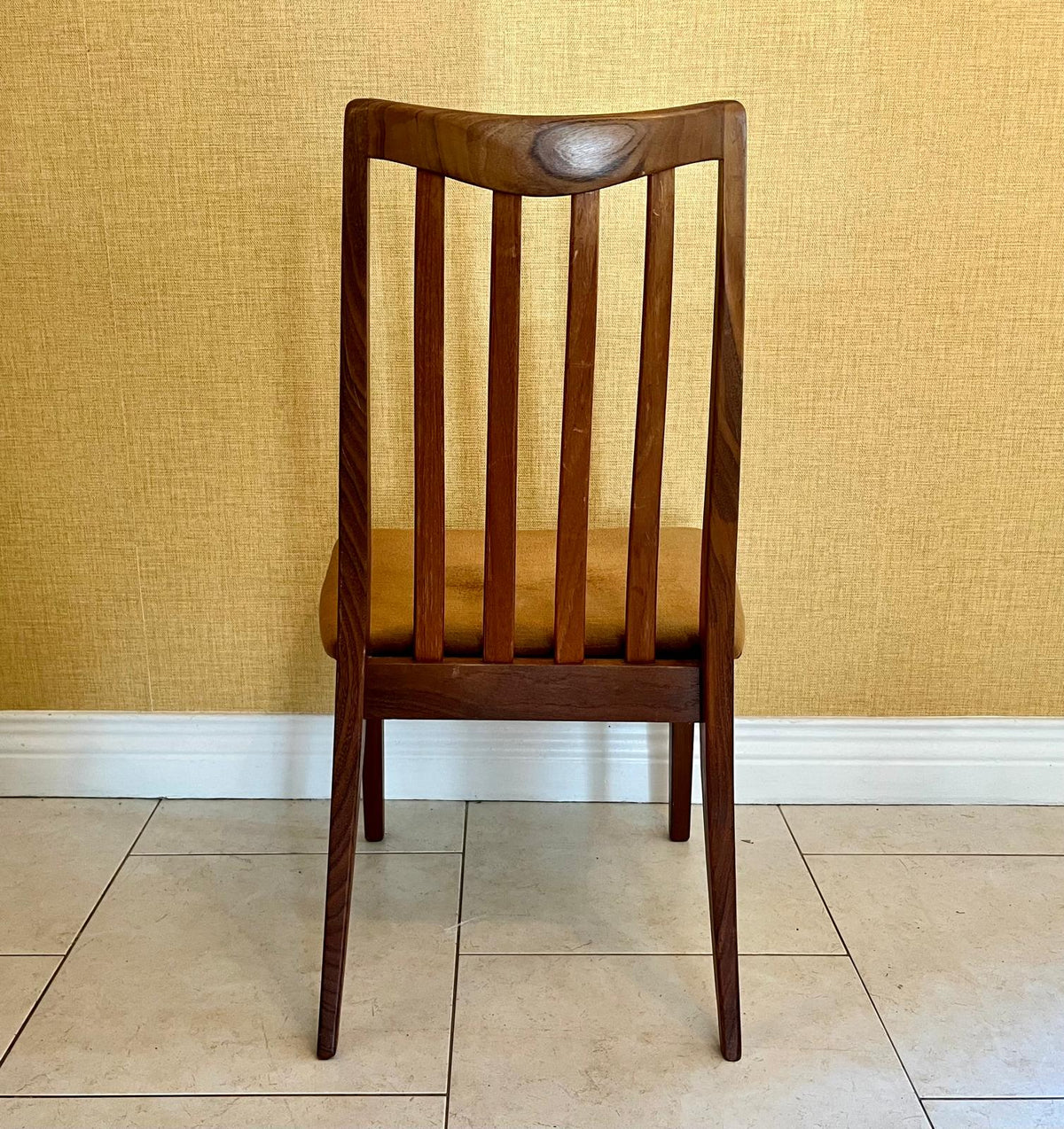 a mid-century G Plan modern wooden dining chair with vertical slat backs and brown velvet seats, arranged on a tiled floor against a textured yellow wall.