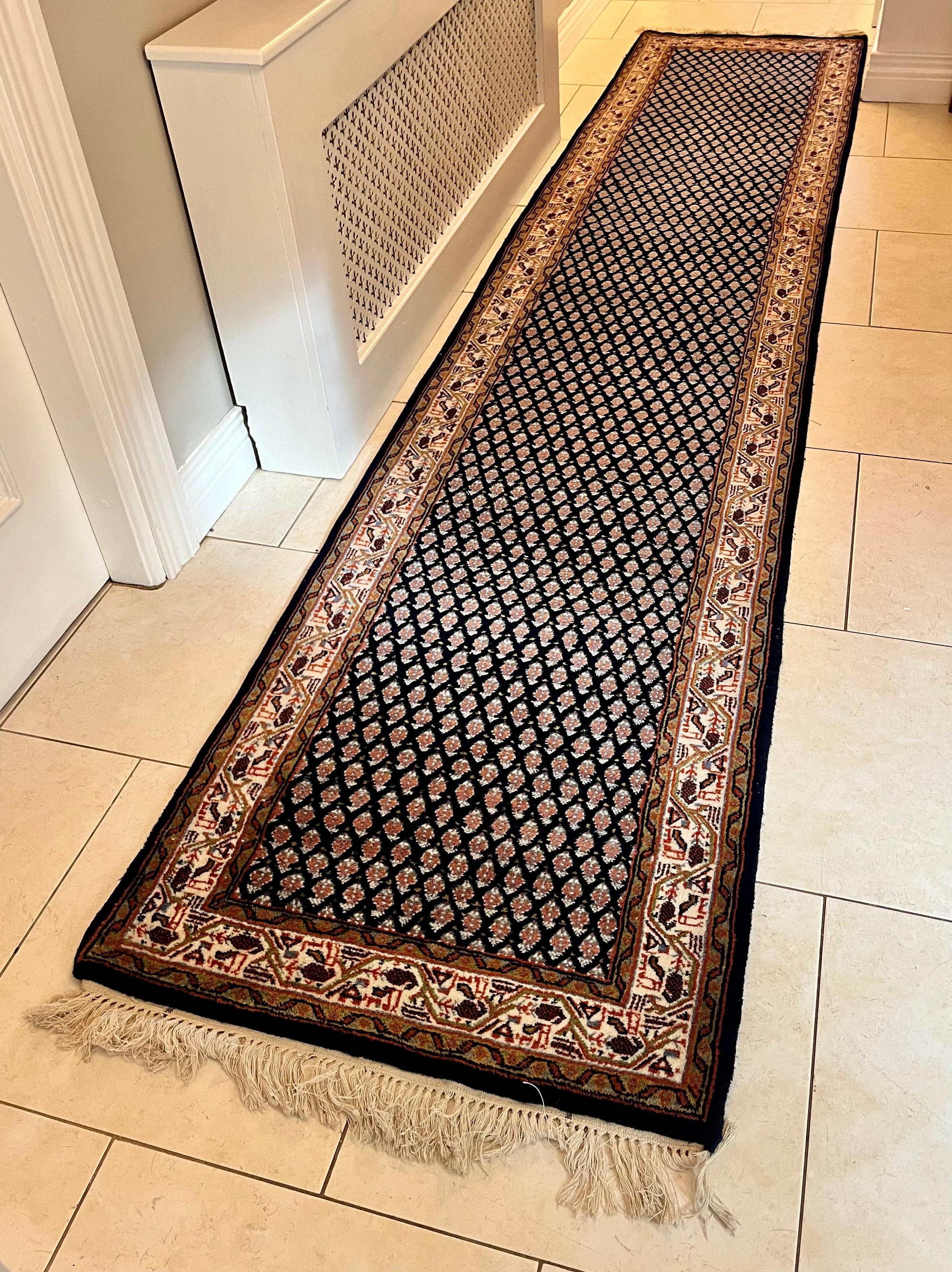 A handmade vintage patterned area rug with a dark navy blue field, cream and orange geometric motifs, and a detailed border, shown laid out on a light-coloured tiled floor with fringe visible at the bottom by Grace & Joy Furniture