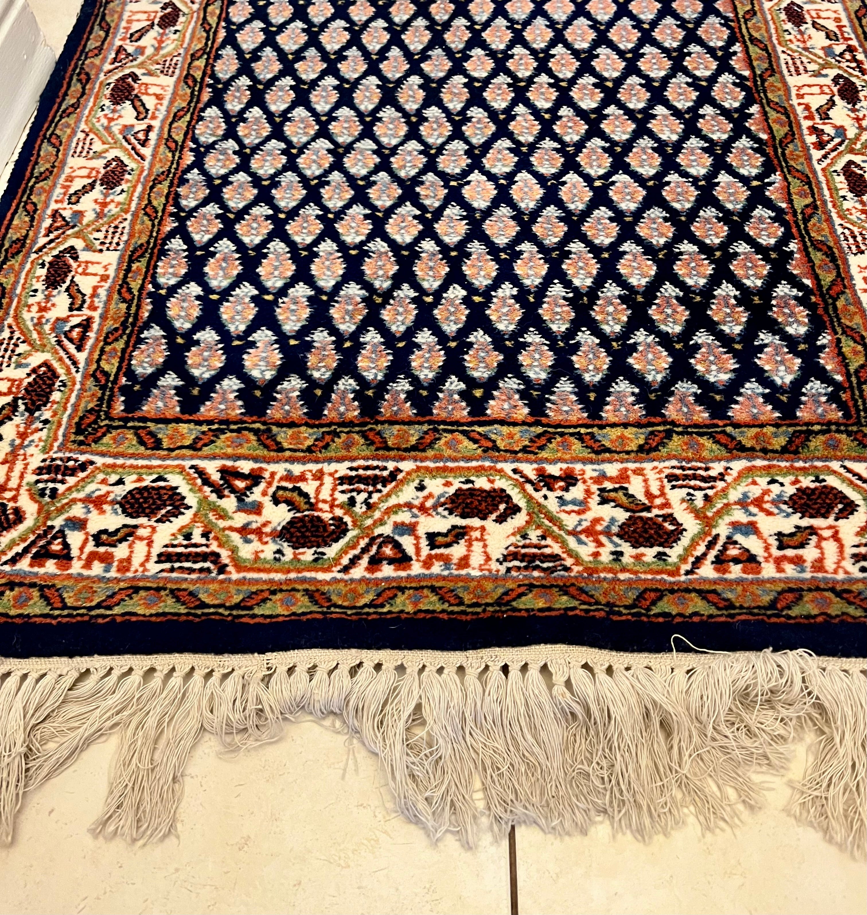 A handmade vintage patterned area rug with a dark navy blue field, cream and orange geometric motifs, and a detailed border, shown laid out on a light-colored tiled floor with fringe visible at the bottom by Grace & Joy Furniture