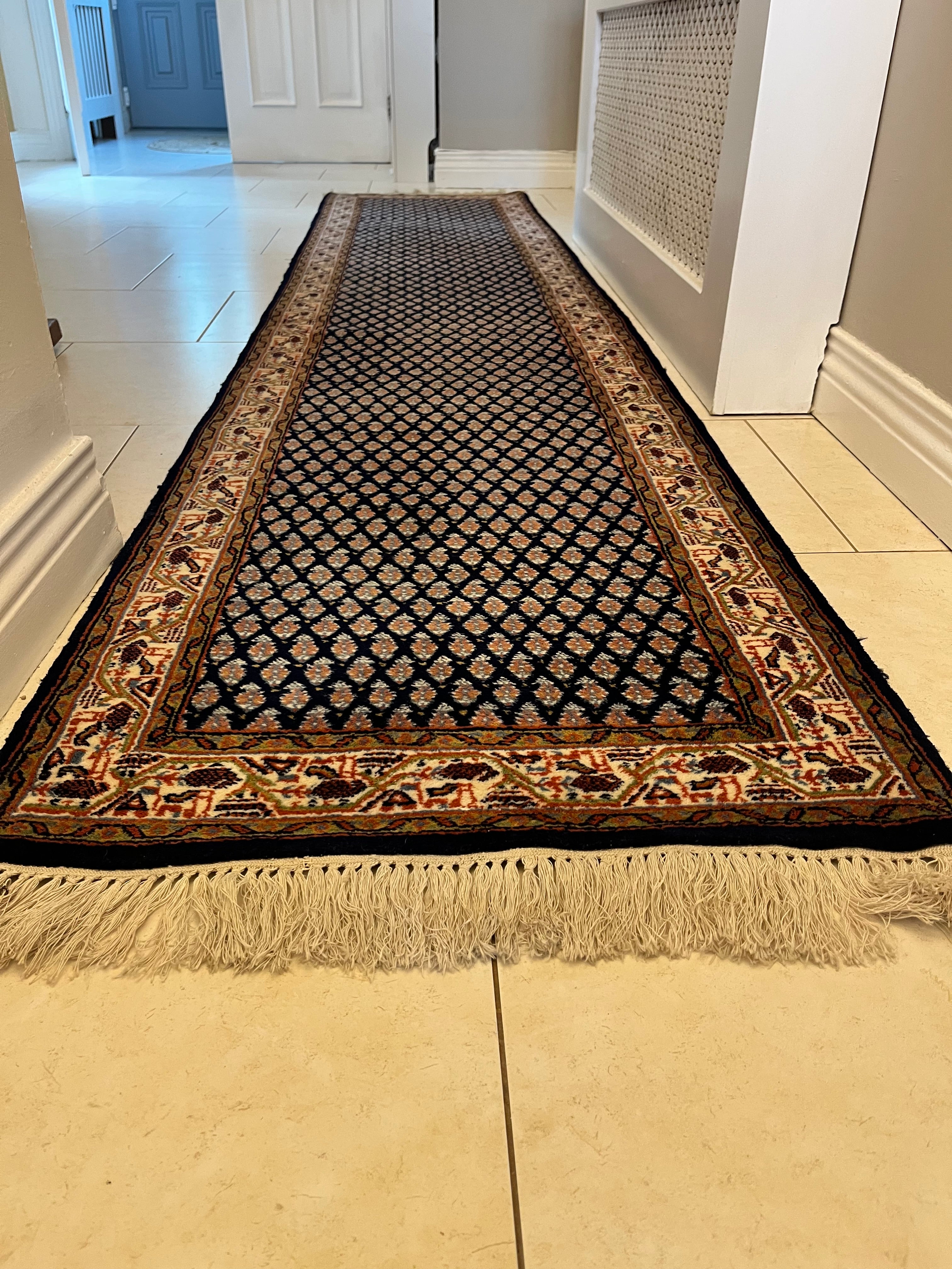 A handmade vintage patterned area rug with a dark navy blue field, cream and orange geometric motifs, and a detailed border, shown laid out on a light-coloured tiled floor with fringe visible at the bottom by Grace & Joy Furniture