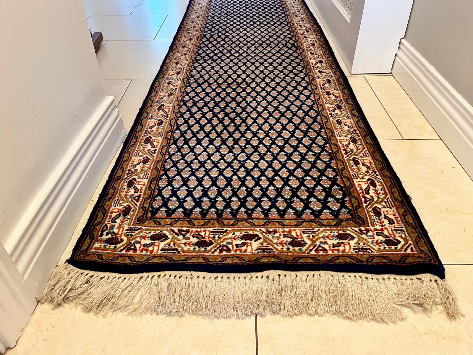 A handmade vintage patterned area rug with a dark navy blue field, cream and orange geometric motifs, and a detailed border, shown laid out on a light-coloured tiled floor with fringe visible at the bottom by Grace & Joy Furniture