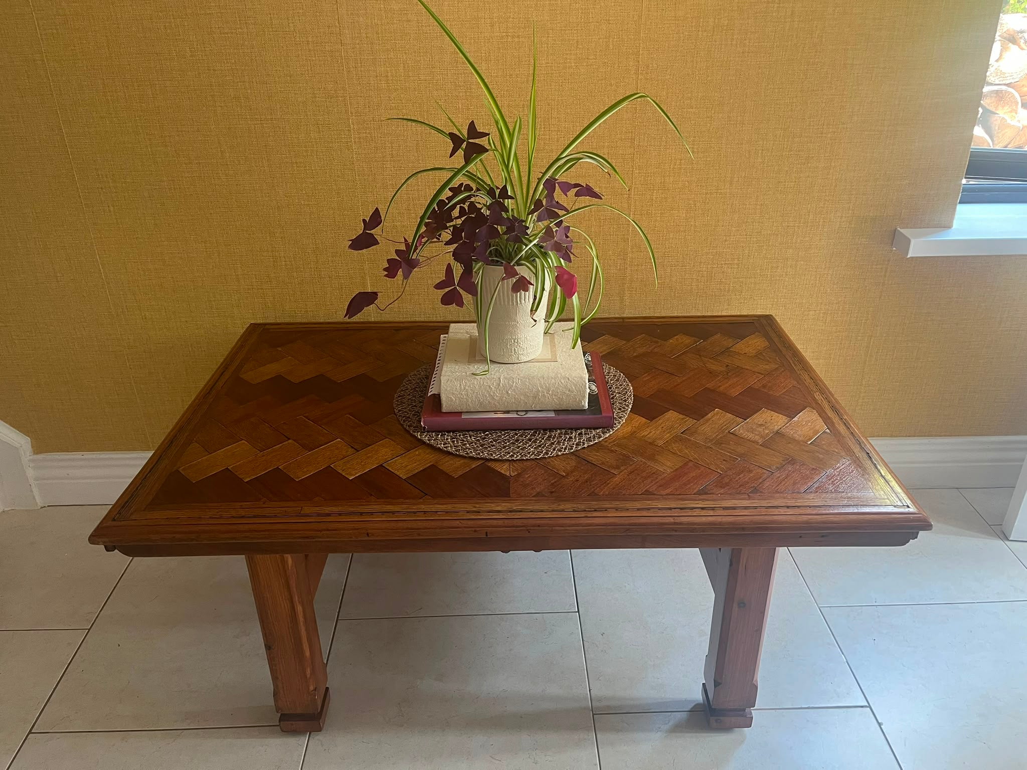 A Herringbone wooden coffee table with a geometric parquet inlay design on the top, set against a textured yellow wall and light-colored tiled floor by Grace & Joy Furniture