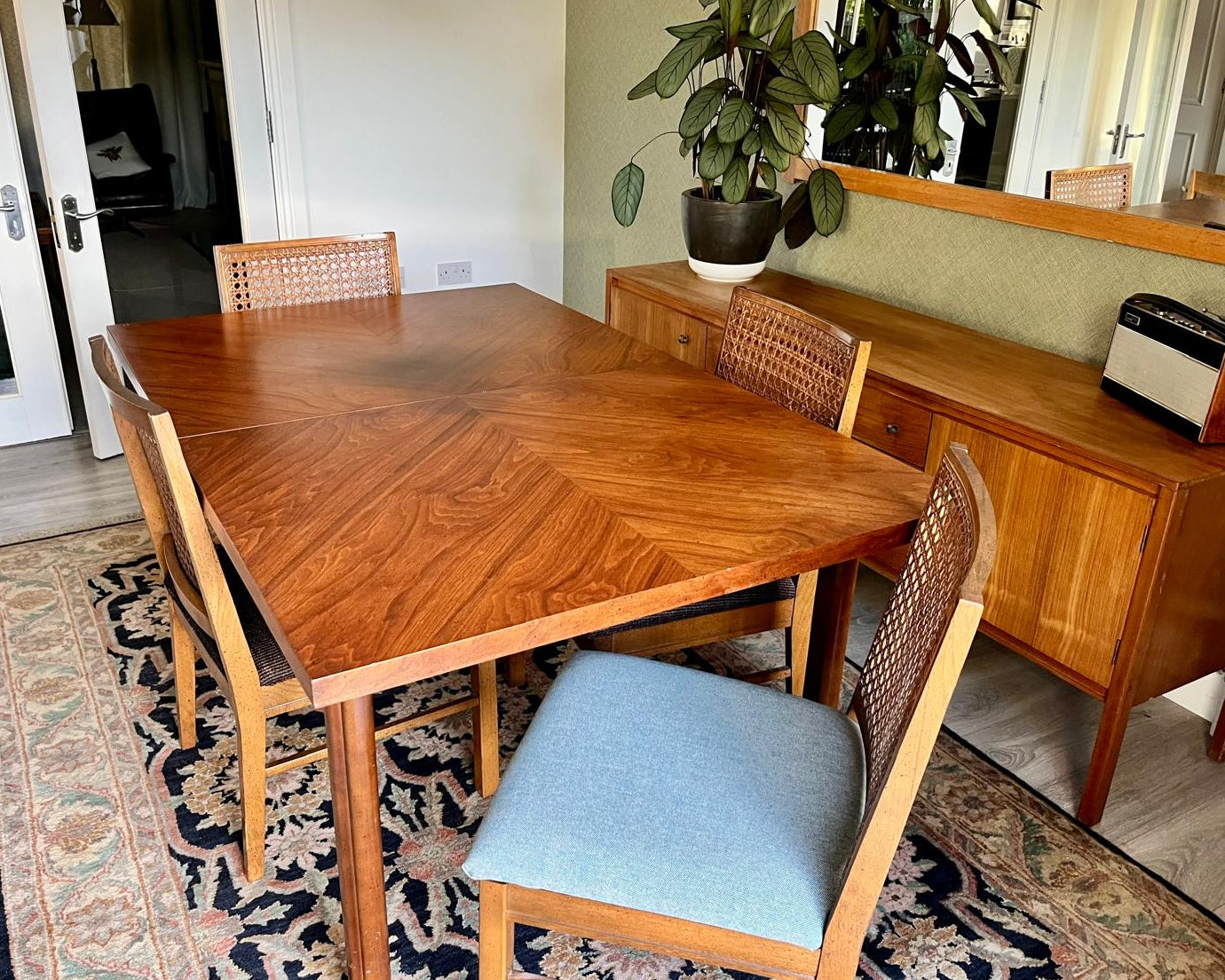A mid-century modern wooden dining table by Lane and four matching chairs with woven backs, set on a patterned area rug in a dining room next to a wooden sideboard.