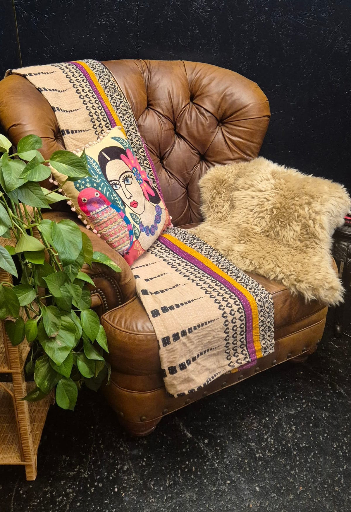 A brown leather armchair in a styled room setting, draped with a beige woven throw blanket featuring black geometric patterns and colorful stripes, alongside a colorful printed pillow and a fluffy beige sheepskin throw. A green potted plant is visible on the left against a dark textured wall.