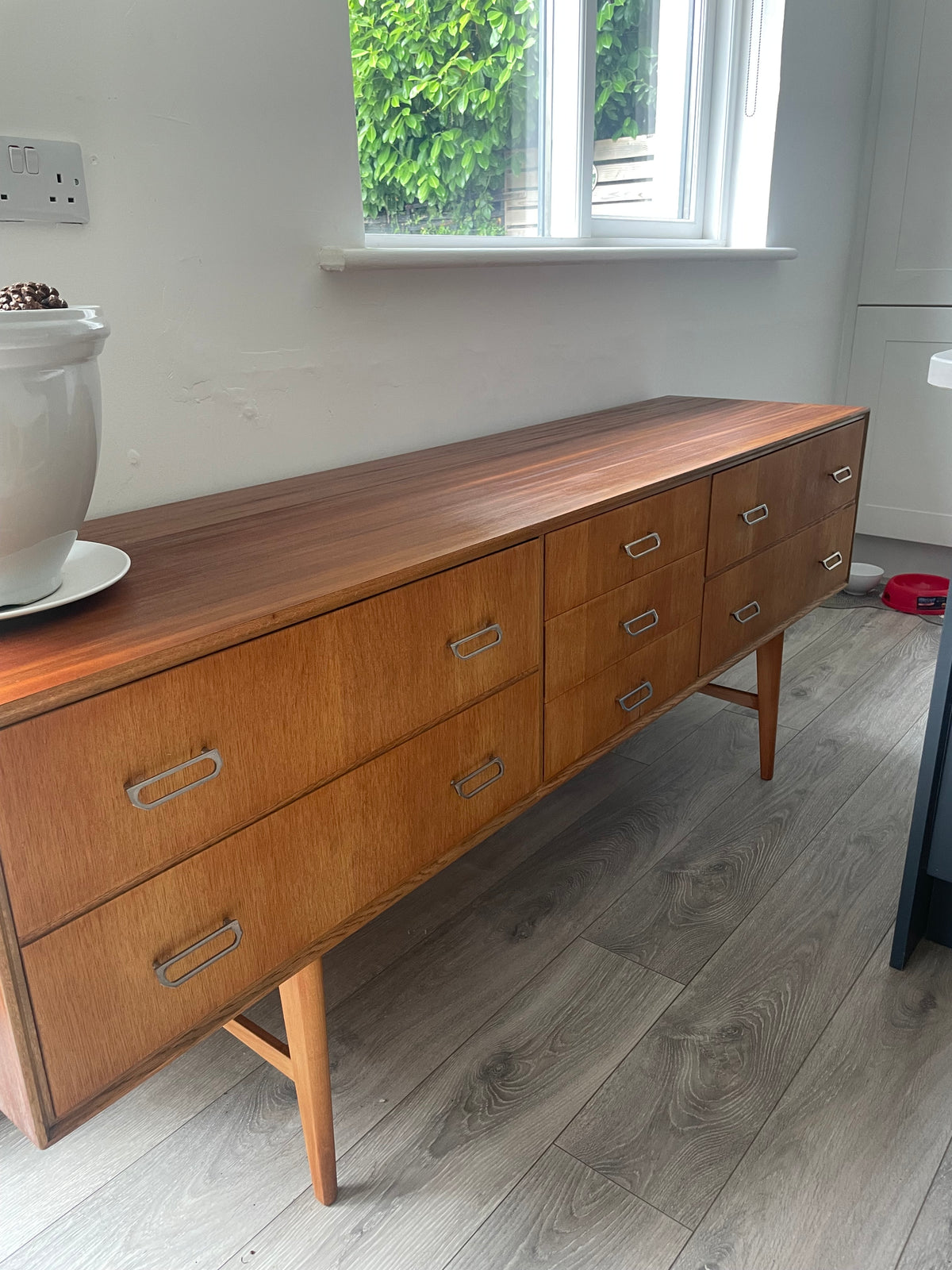 Mid-century modern wooden meredew credenza with six drawers and silver handles, shown in a styled room with gray wood floors, a green wall, and a mirror behind it.