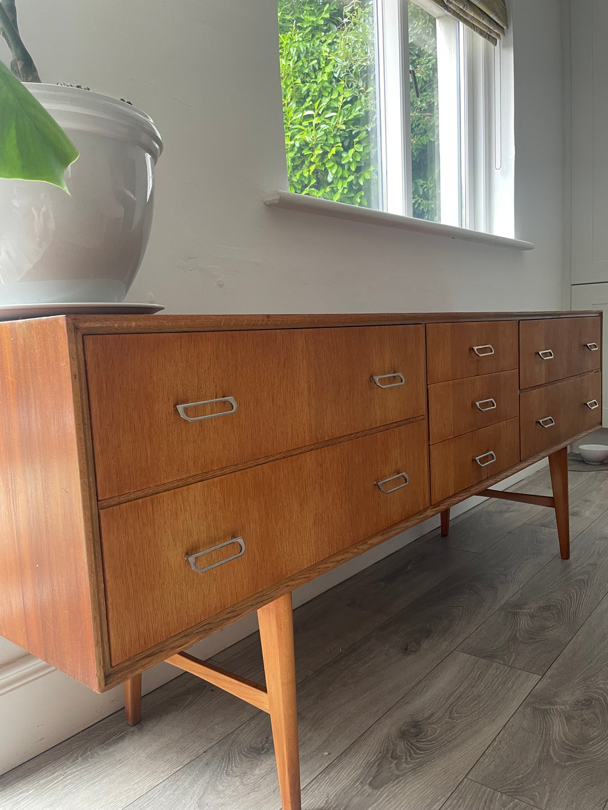Mid-century modern wooden meredew credenza with six drawers and silver handles, shown in a styled room with gray wood floors, a green wall, and a mirror behind it.