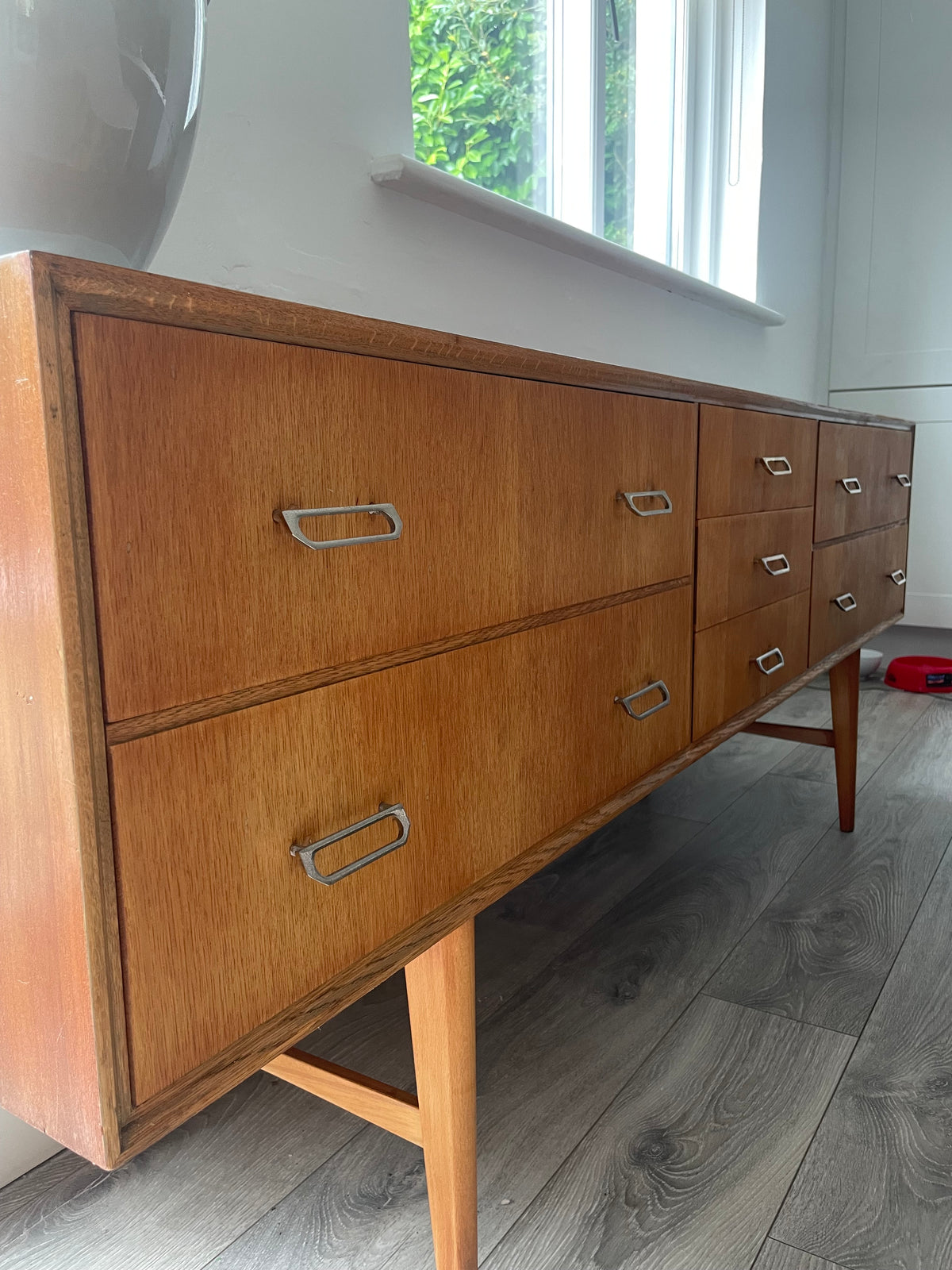 Mid-century modern wooden meredew credenza with six drawers and silver handles, shown in a styled room with gray wood floors, a green wall, and a mirror behind it.