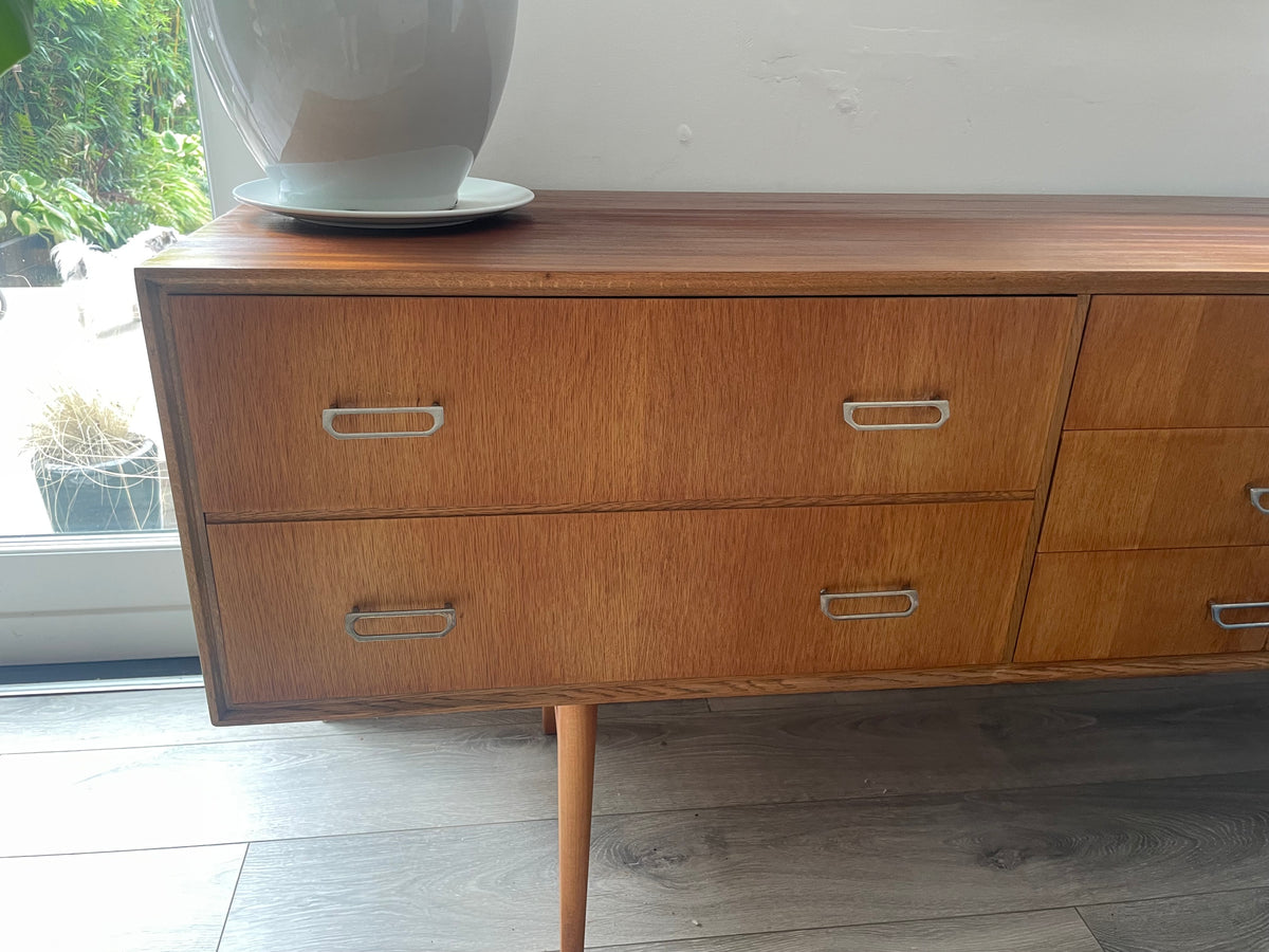 Mid-century modern wooden meredew credenza with six drawers and silver handles, shown in a styled room with gray wood floors, a green wall, and a mirror behind it.