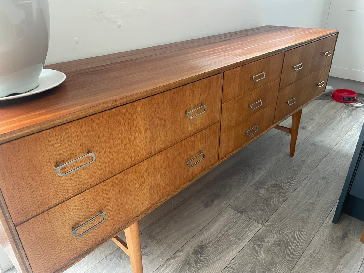 Mid-century modern wooden meredew credenza with six drawers and silver handles, shown in a styled room with gray wood floors, a green wall, and a mirror behind it.