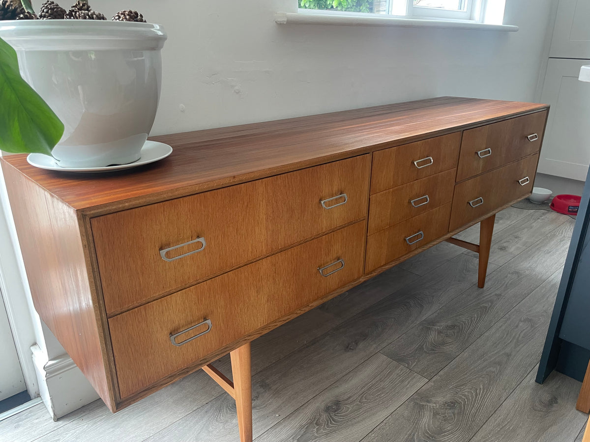 Mid-century modern wooden meredew credenza with six drawers and silver handles, shown in a styled room with gray wood floors, a green wall, and a mirror behind it.
