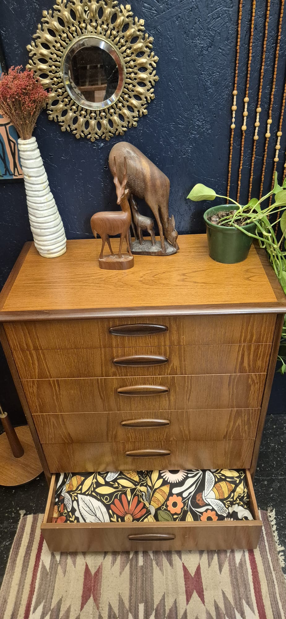 A mid-century modern wooden chest of drawers with five drawers, shown with the bottom drawer open to reveal a floral patterned interior lining. The dresser is styled with decorative items including a small elephant figurine, a striped vase, and a potted plant against a dark blue textured wall.