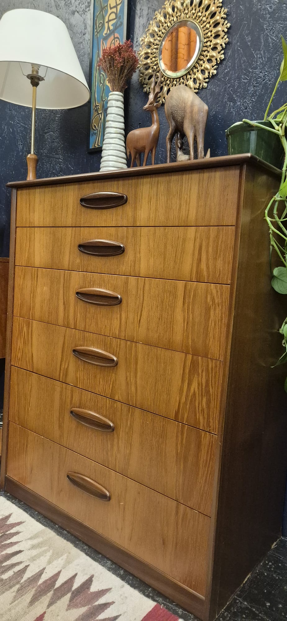 A mid-century modern wooden chest of drawers with six drawers and dark wood handles, styled in a room setting next to a lamp and decorative items.