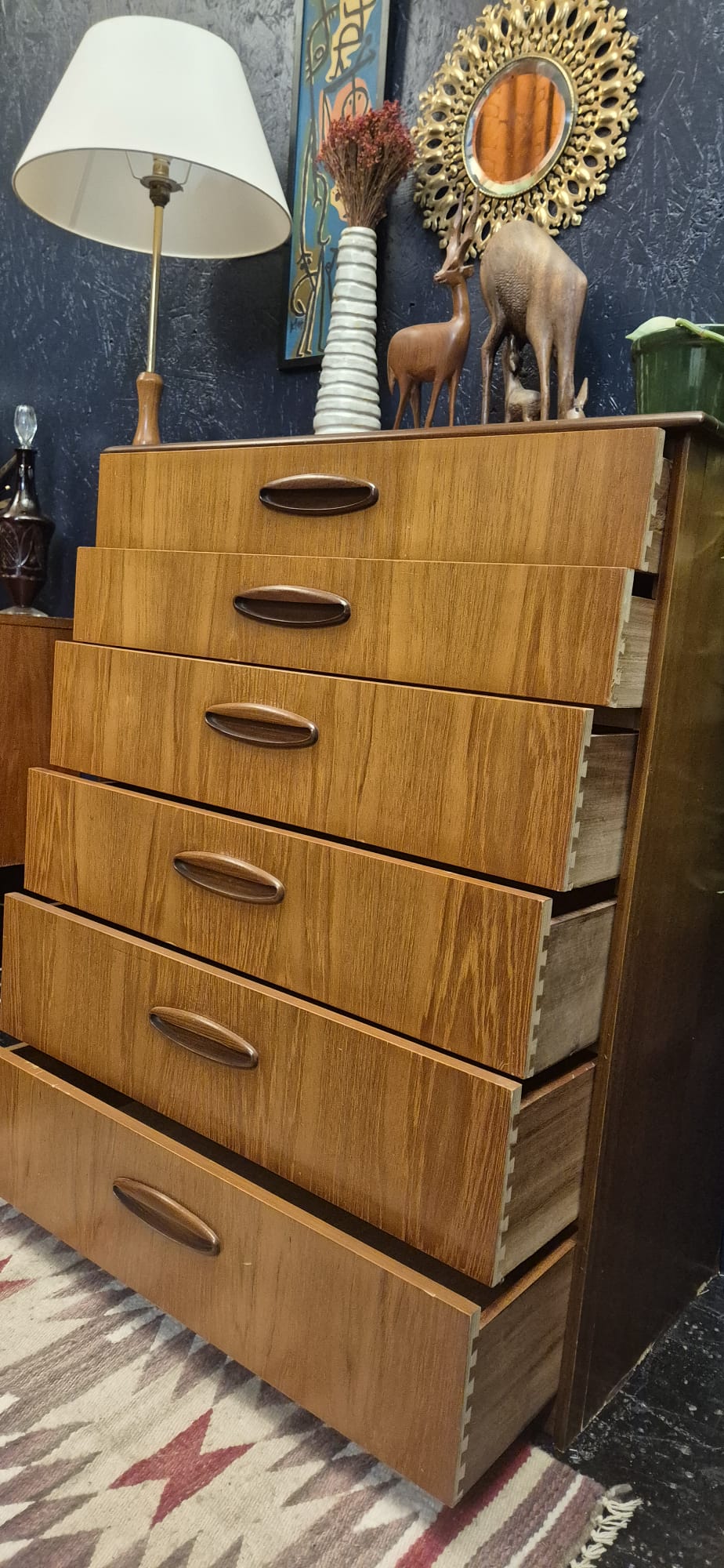 A mid-century modern wooden chest of drawers with six drawers and curved metal handles, styled in a home interior next to a lamp and decorative items.
