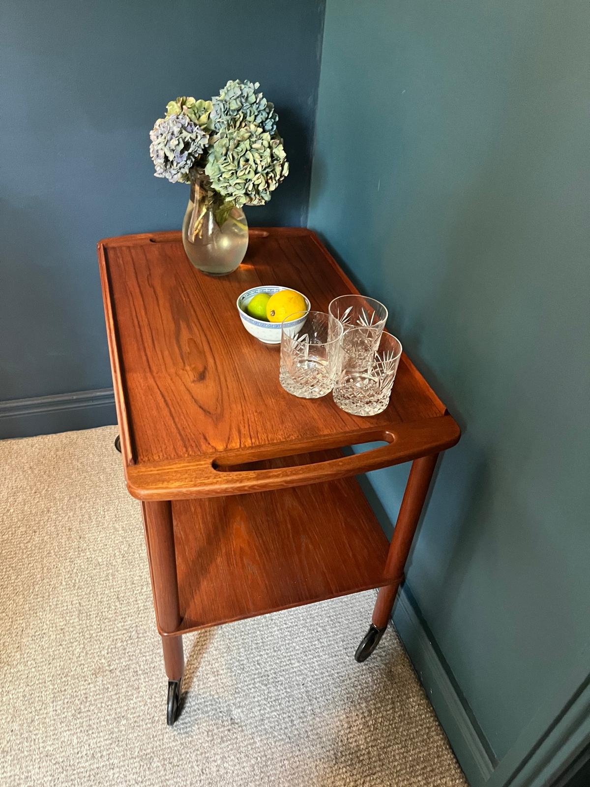 A mid-century teak Bowa drinks trolley, styled with a glass vase of hydrangeas, a small bowl of lemons, and two cut-crystal glasses against a dark teal wall and beige carpet by Grace & Joy Furniture