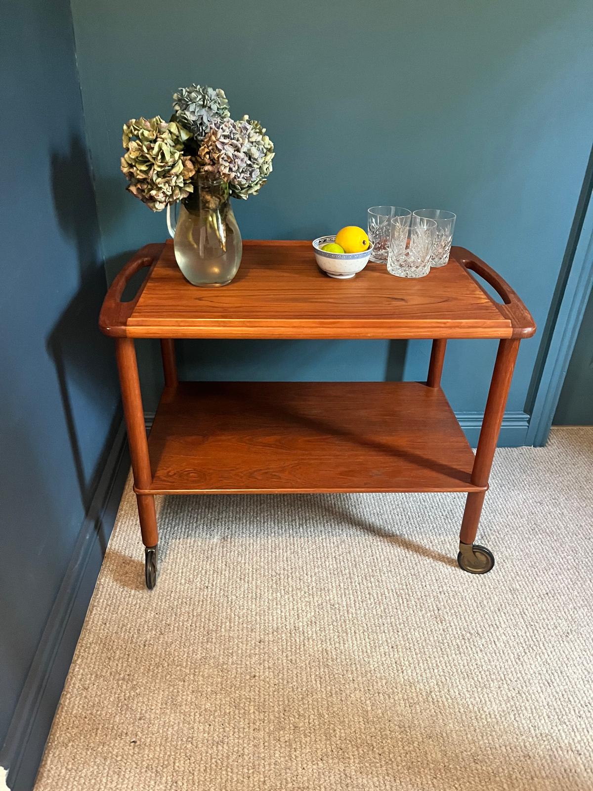A mid-century teak Bowa drinks trolley, styled with a glass vase of hydrangeas, a small bowl of lemons, and two cut-crystal glasses against a dark teal wall and beige carpet by Grace & Joy Furniture