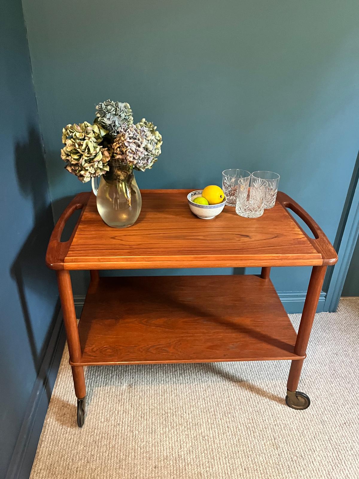 A mid-century teak Bowa drinks trolley, styled with a glass vase of hydrangeas, a small bowl of lemons, and two cut-crystal glasses against a dark teal wall and beige carpet by Grace & Joy Furniture