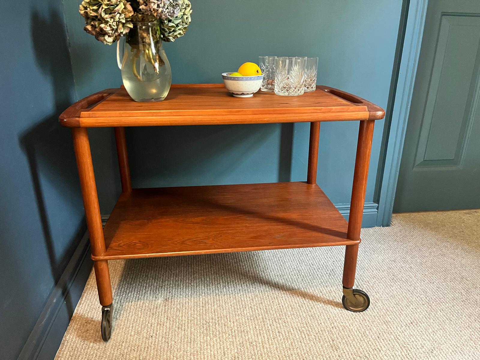 A mid-century teak Bowa drinks trolley, styled with a glass vase of hydrangeas, a small bowl of lemons, and two cut-crystal glasses against a dark teal wall and beige carpet by Grace & Joy Furniture