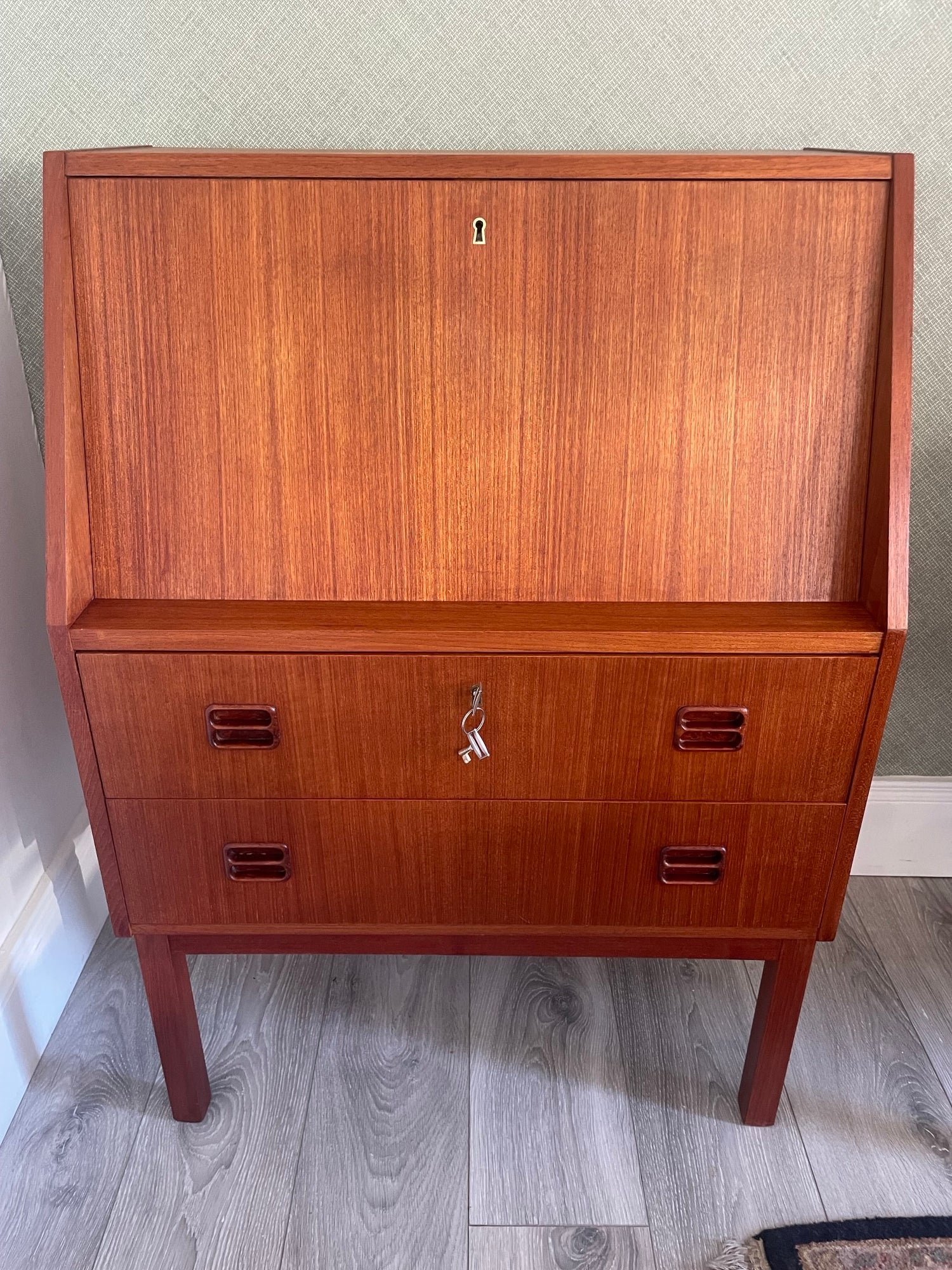 A mid-century modern Danish bureau with a warm reddish-brown finish, featuring a large left door and three drawers on the right with dark metal pulls, photographed indoors on a gray wood floor. by Grace & Joy Furniture