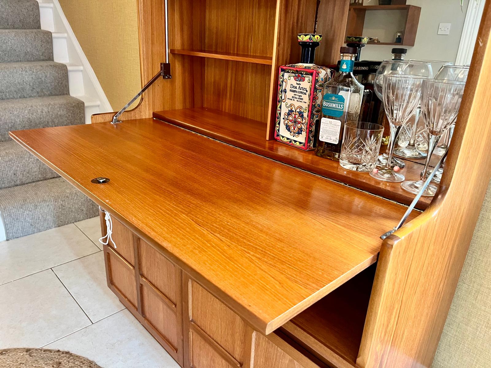 A Nathan mid century drinks cabinet with a drop-down wooden countertop, shown in a home setting next to a staircase. The cabinet is styled with liquor bottles, crystal glasses, and a small shelf inside by Grace & Joy Furniture