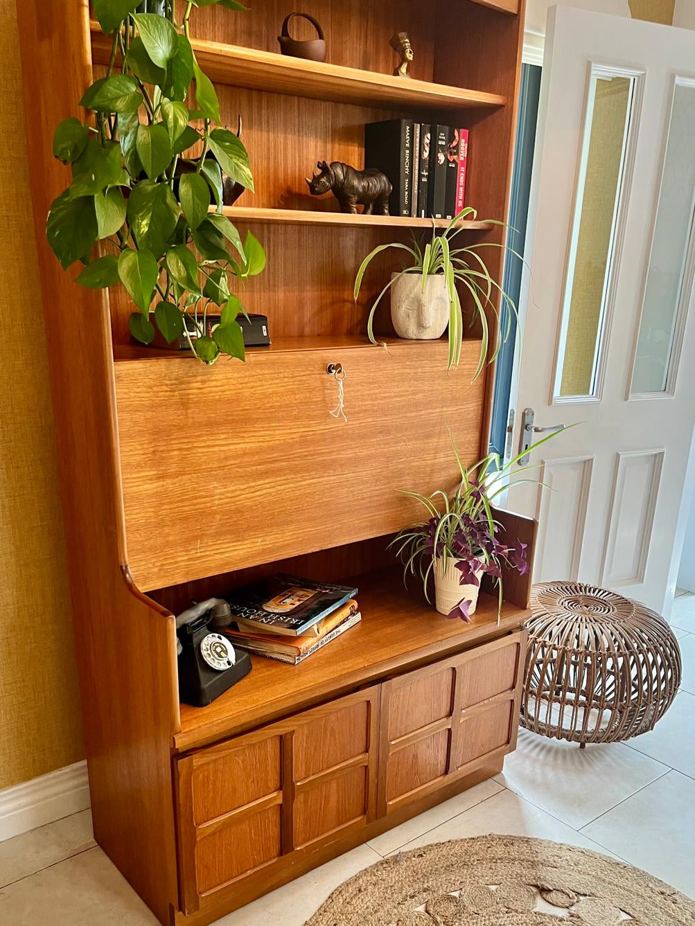 A Nathan mid century drinks cabinet with a drop-down wooden countertop, shown in a home setting next to a staircase. The cabinet is styled with liquor bottles, crystal glasses, and a small shelf inside by Grace & Joy Furniture