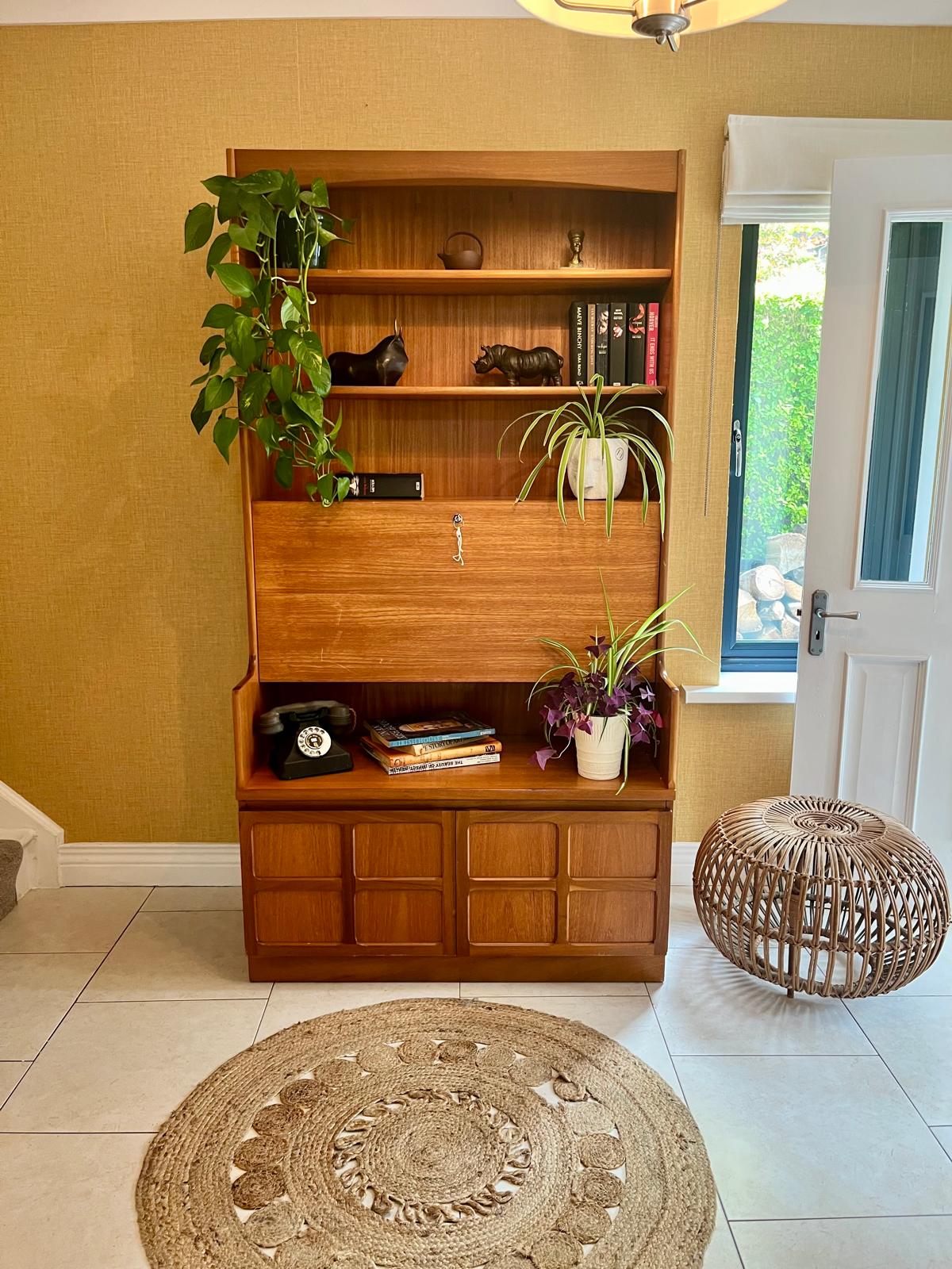 A Nathan mid century drinks cabinet with a drop-down wooden countertop, shown in a home setting next to a staircase. The cabinet is styled with liquor bottles, crystal glasses, and a small shelf inside by Grace & Joy Furniture