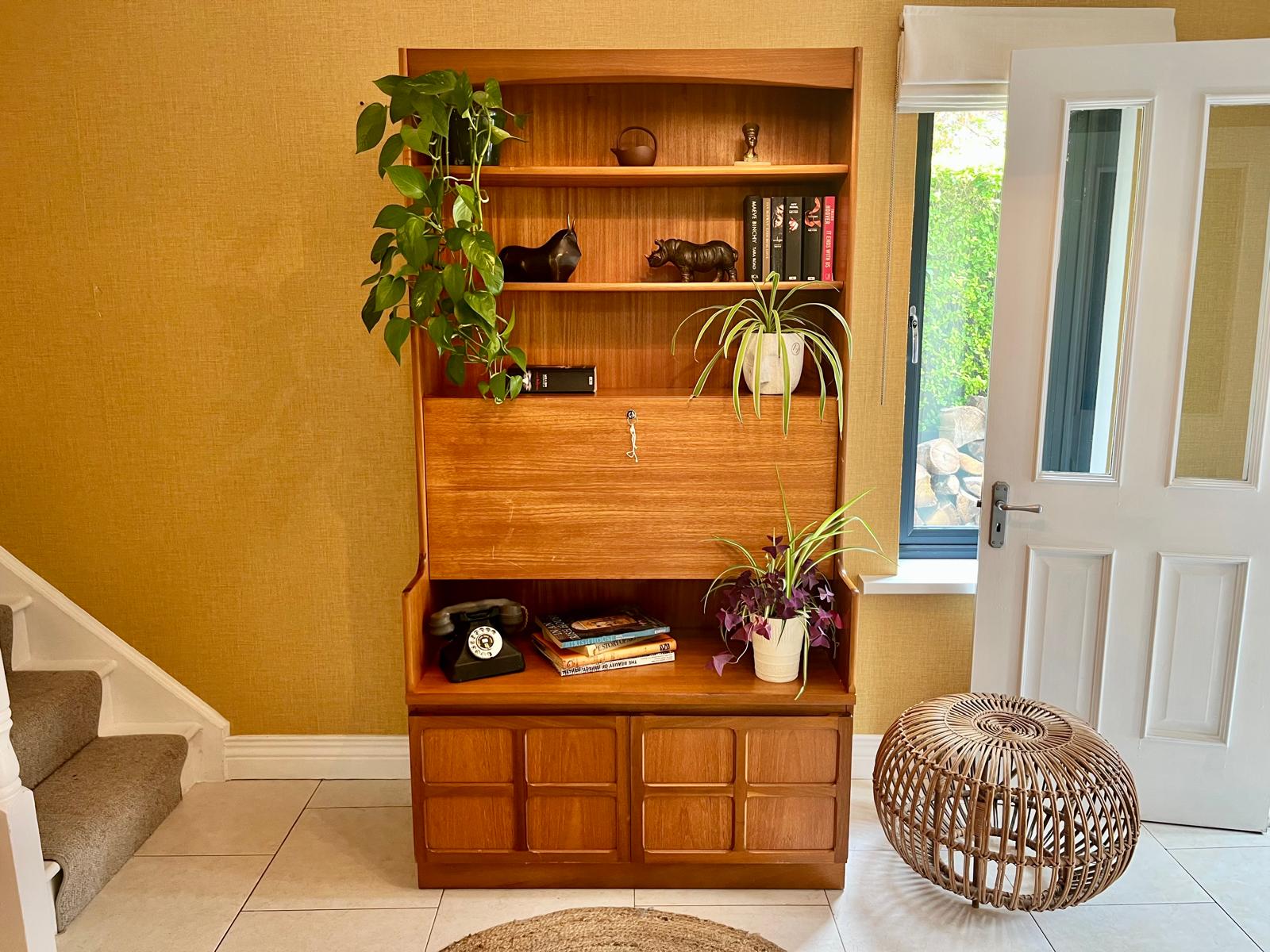 A Nathan mid century drinks cabinet with a drop-down wooden countertop, shown in a home setting next to a staircase. The cabinet is styled with liquor bottles, crystal glasses, and a small shelf inside by Grace & Joy Furniture