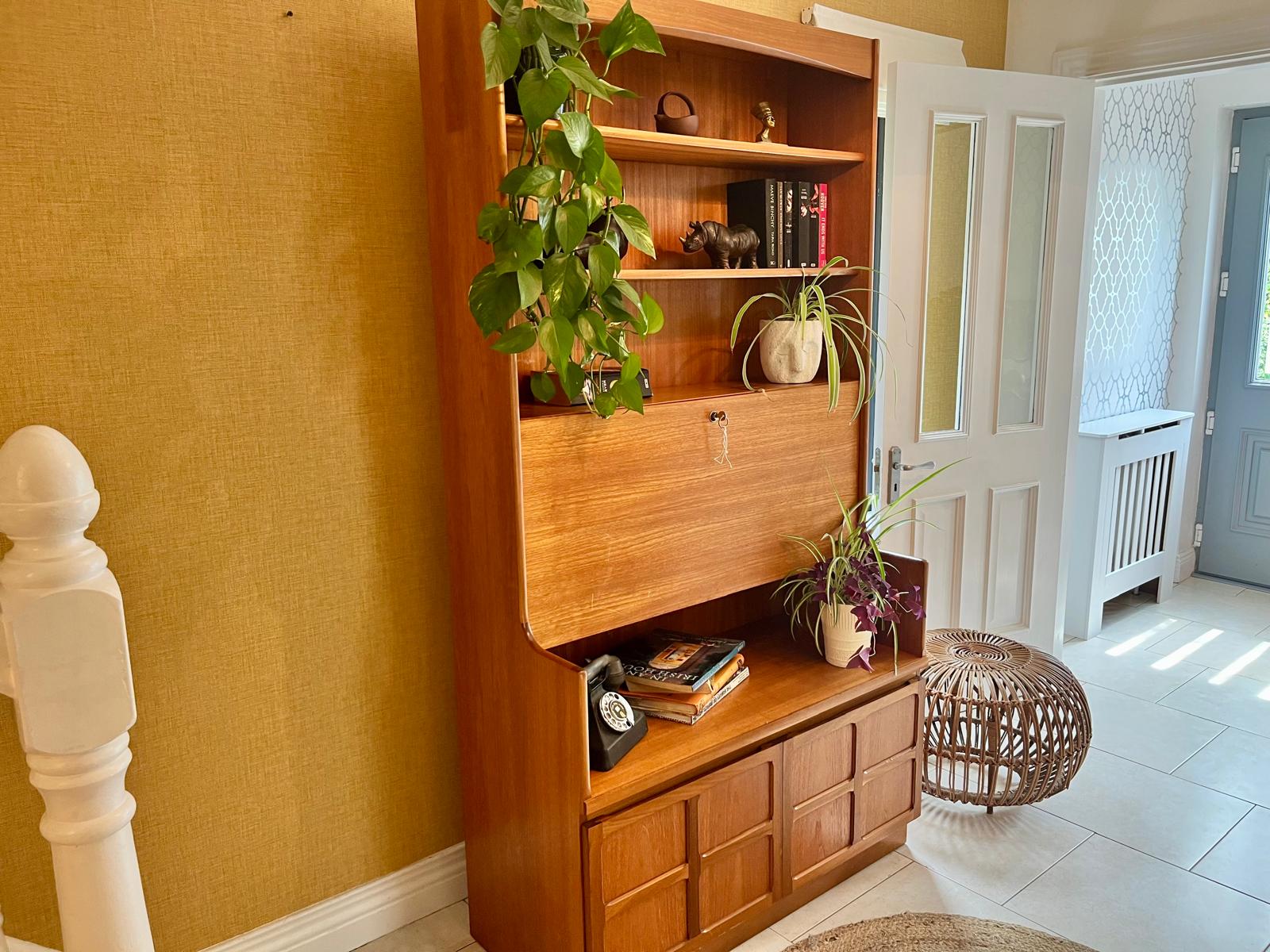 A Nathan mid century drinks cabinet with a drop-down wooden countertop, shown in a home setting next to a staircase. The cabinet is styled with liquor bottles, crystal glasses, and a small shelf inside by Grace & Joy Furniture