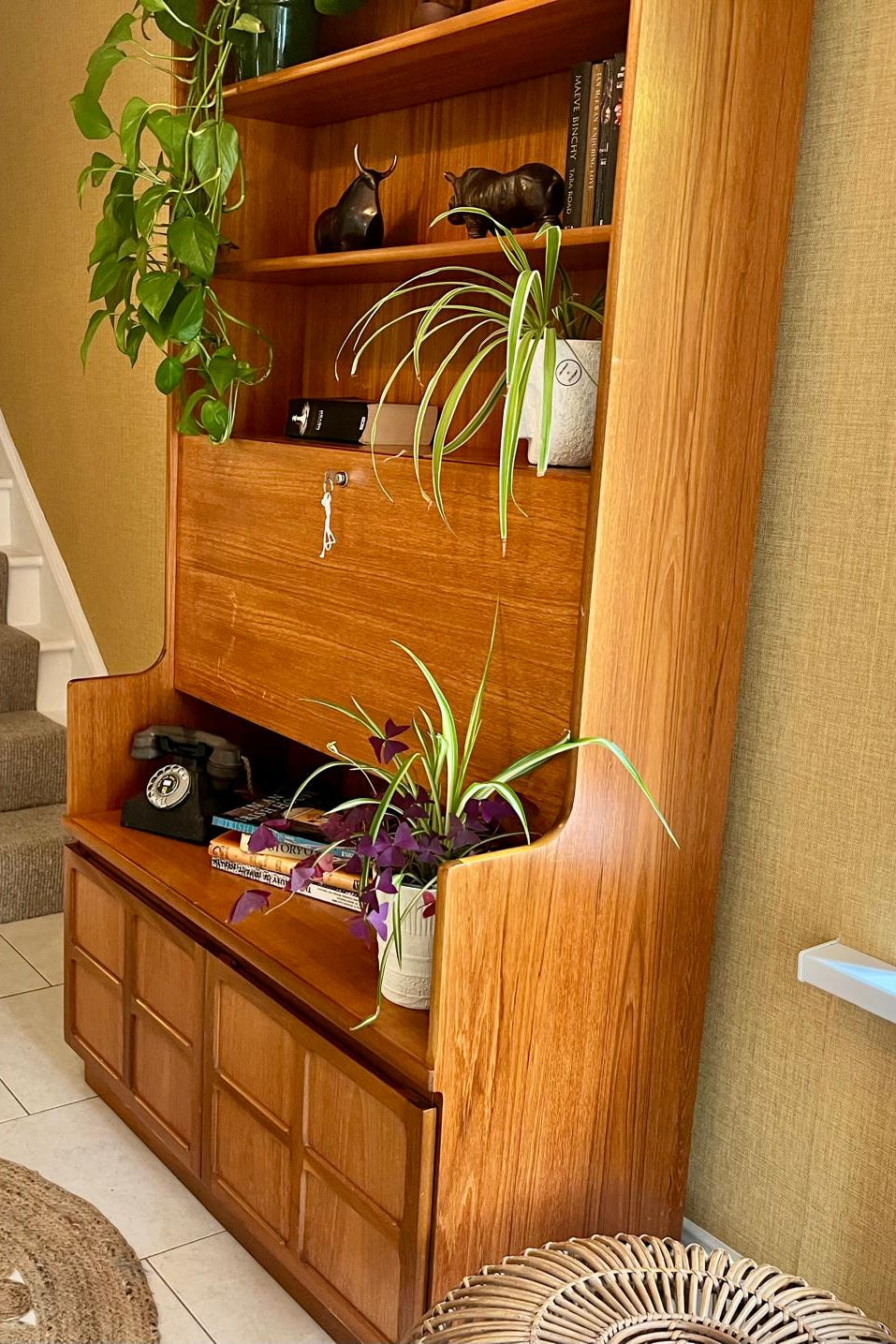 A Nathan mid century drinks cabinet with a drop-down wooden countertop, shown in a home setting next to a staircase. The cabinet is styled with liquor bottles, crystal glasses, and a small shelf inside by Grace & Joy Furniture