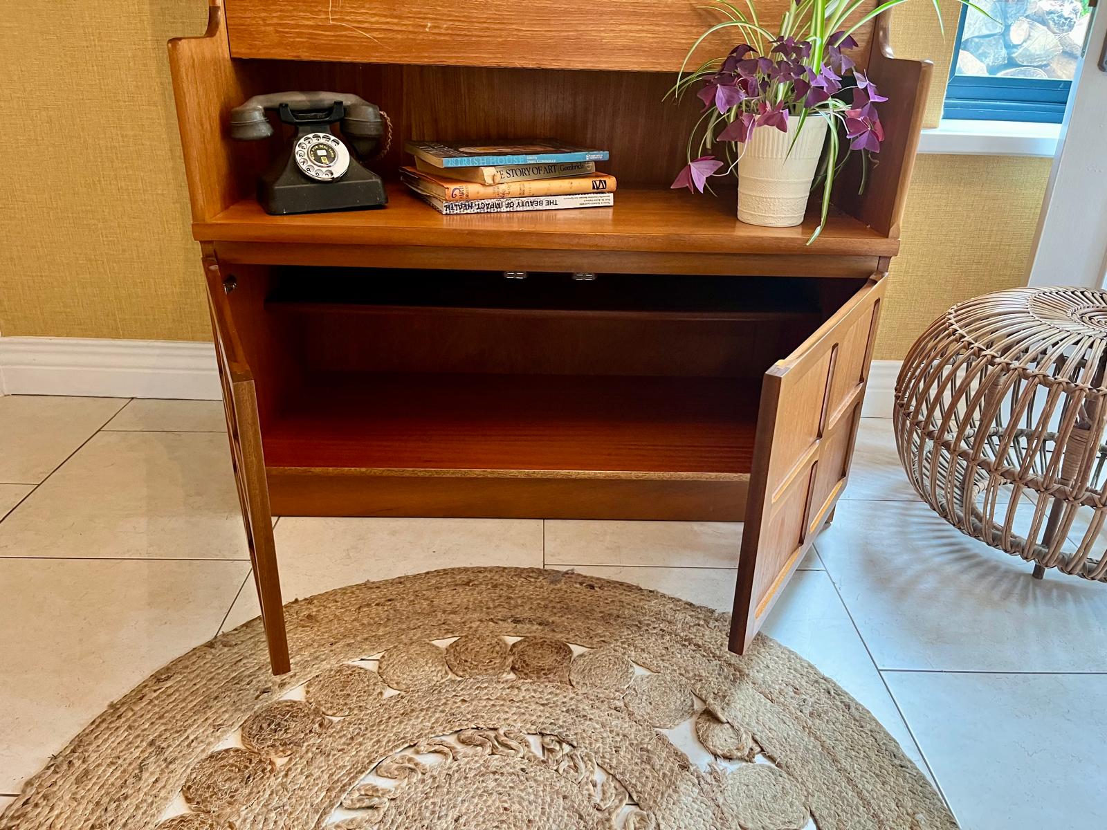 A Nathan mid century drinks cabinet with a drop-down wooden countertop, shown in a home setting next to a staircase. The cabinet is styled with liquor bottles, crystal glasses, and a small shelf inside by Grace & Joy Furniture