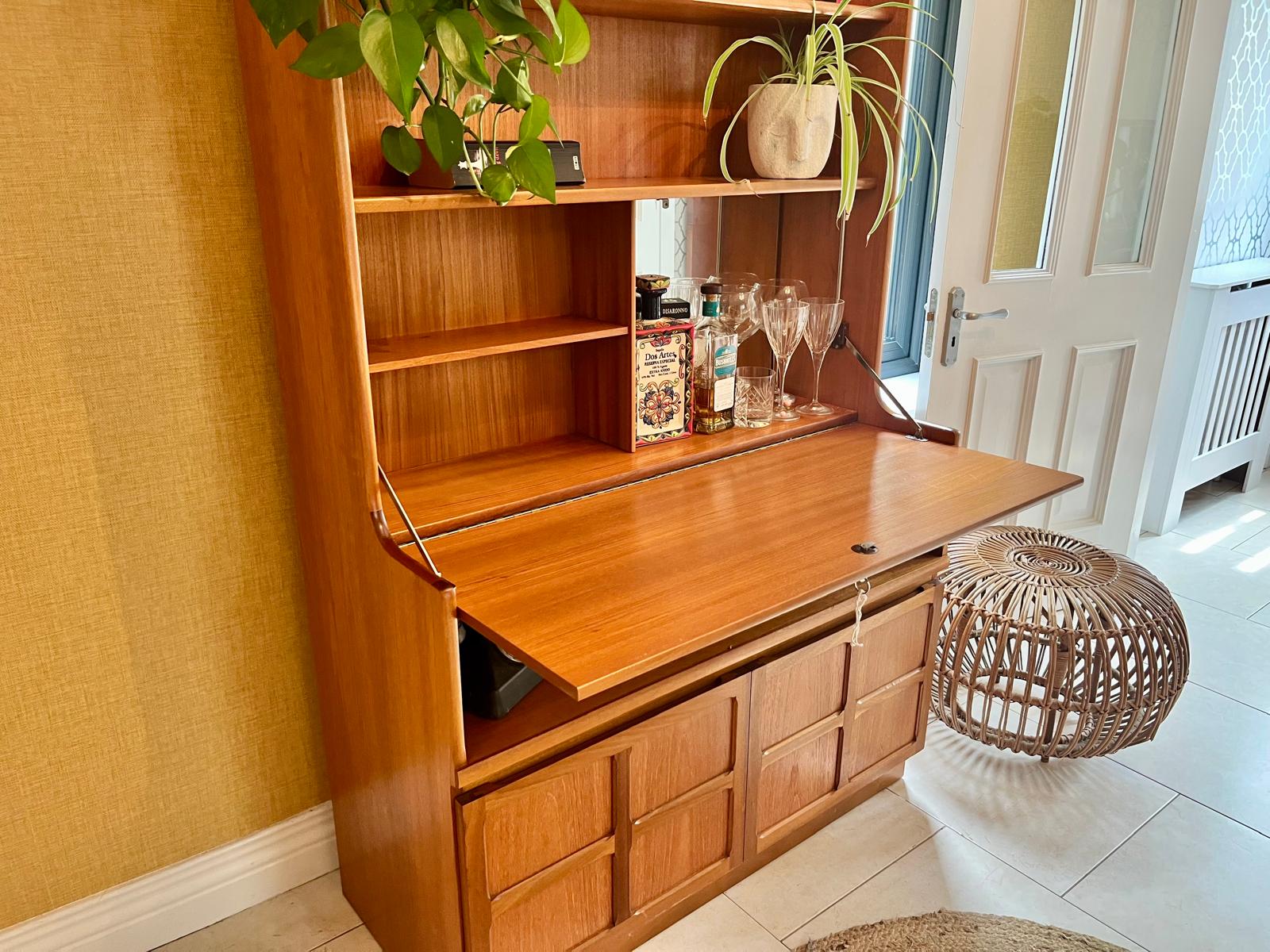 A Nathan mid century drinks cabinet with a drop-down wooden countertop, shown in a home setting next to a staircase. The cabinet is styled with liquor bottles, crystal glasses, and a small shelf inside by Grace & Joy Furniture