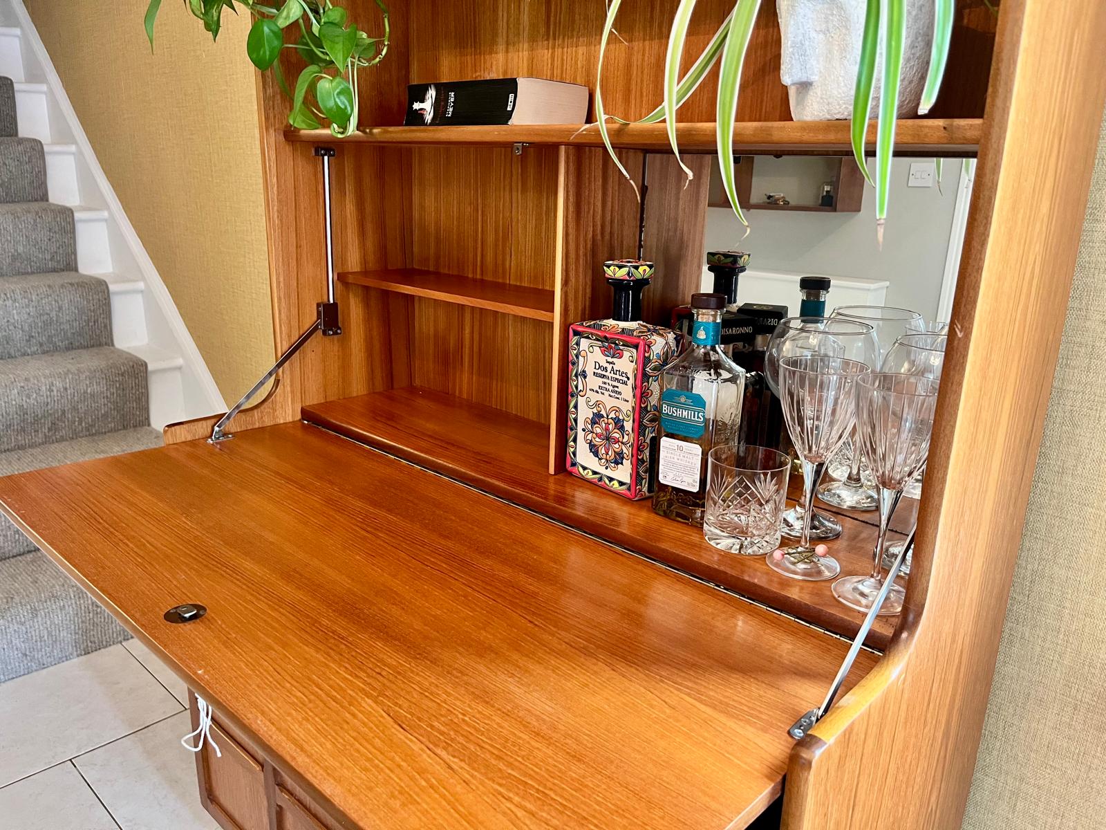 A Nathan mid century drinks cabinet with a drop-down wooden countertop, shown in a home setting next to a staircase. The cabinet is styled with liquor bottles, crystal glasses, and a small shelf inside by Grace & Joy Furniture