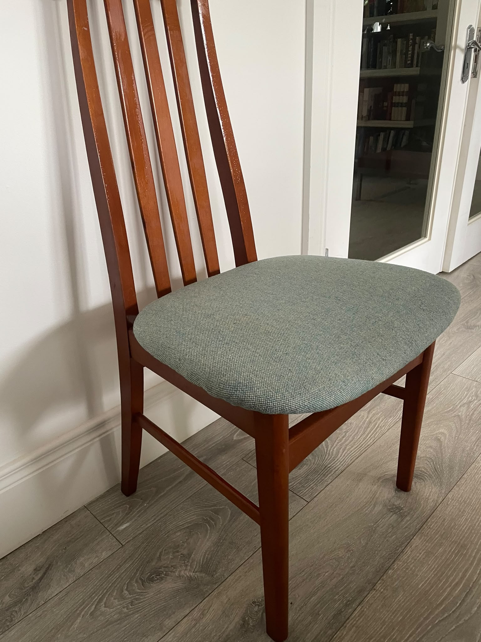 A wooden mid century Jenitique dining chair with a tall slatted backrest and a light blue upholstered seat cushion, shown on a gray wood floor against a white wall by Grace & Joy Furniture