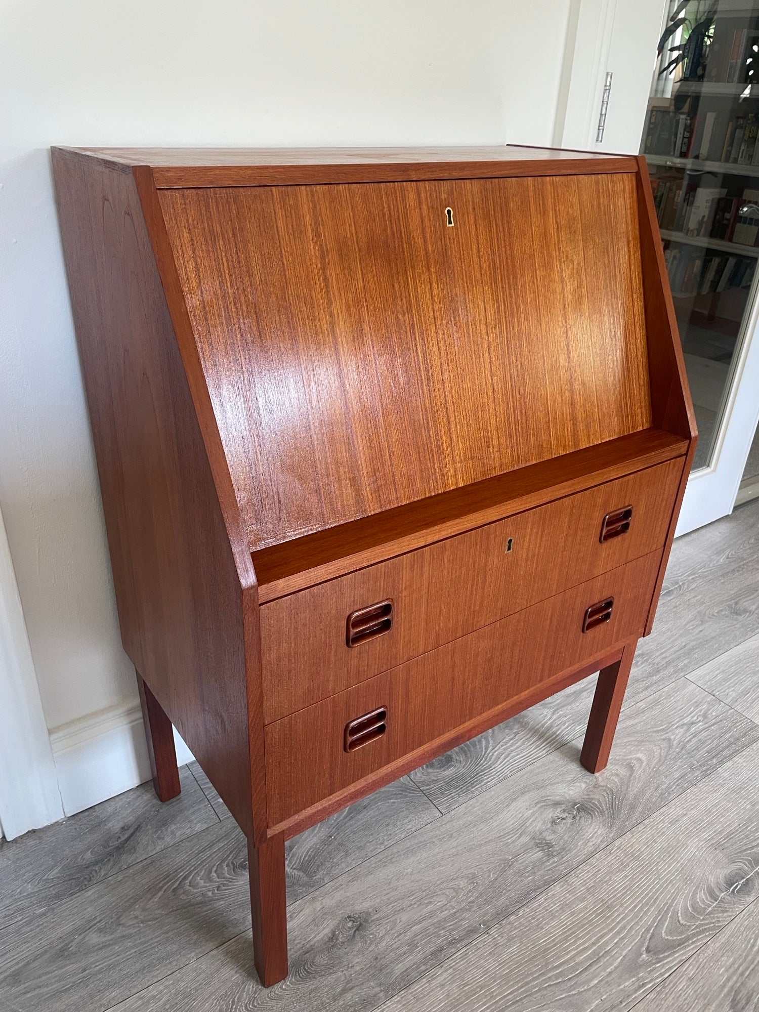 A mid-century modern Danish bureau with a warm reddish-brown finish, featuring a large left door and three drawers on the right with dark metal pulls, photographed indoors on a gray wood floor. by Grace & Joy Furniture