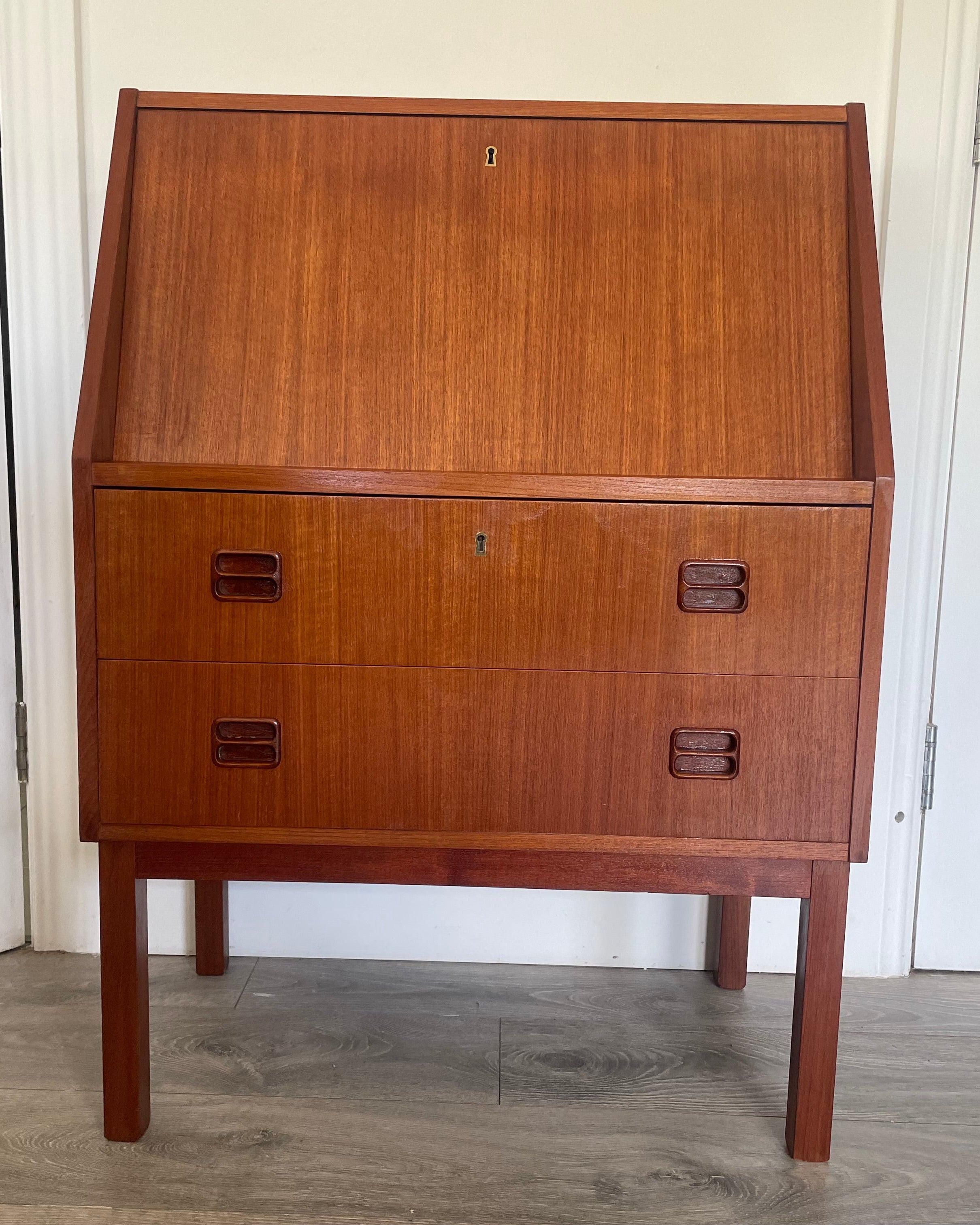 A mid-century modern Danish bureau with a warm reddish-brown finish, featuring a large left door and three drawers on the right with dark metal pulls, photographed indoors on a gray wood floor. by Grace & Joy Furniture
