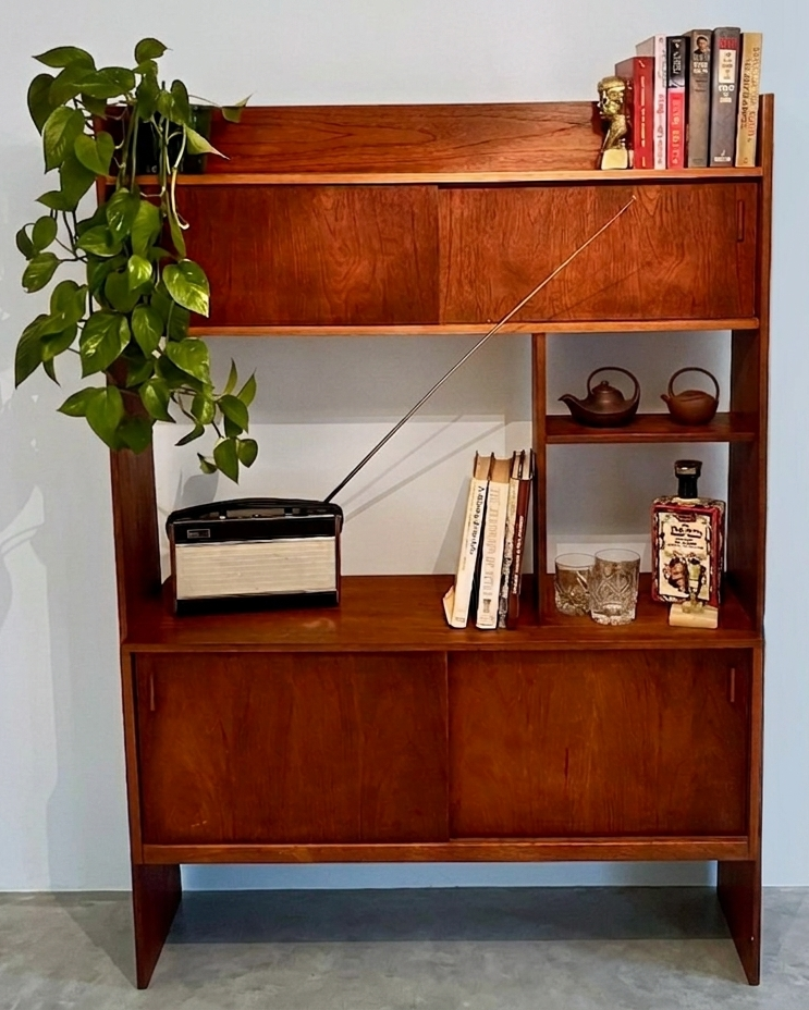 Mid-century modern wooden room divider with a warm cherry finish, featuring a top cabinet, open shelves, and lower cabinet doors, styled with books, a radio, and a trailing green plant against a light blue wall by Grace & Joy furniture