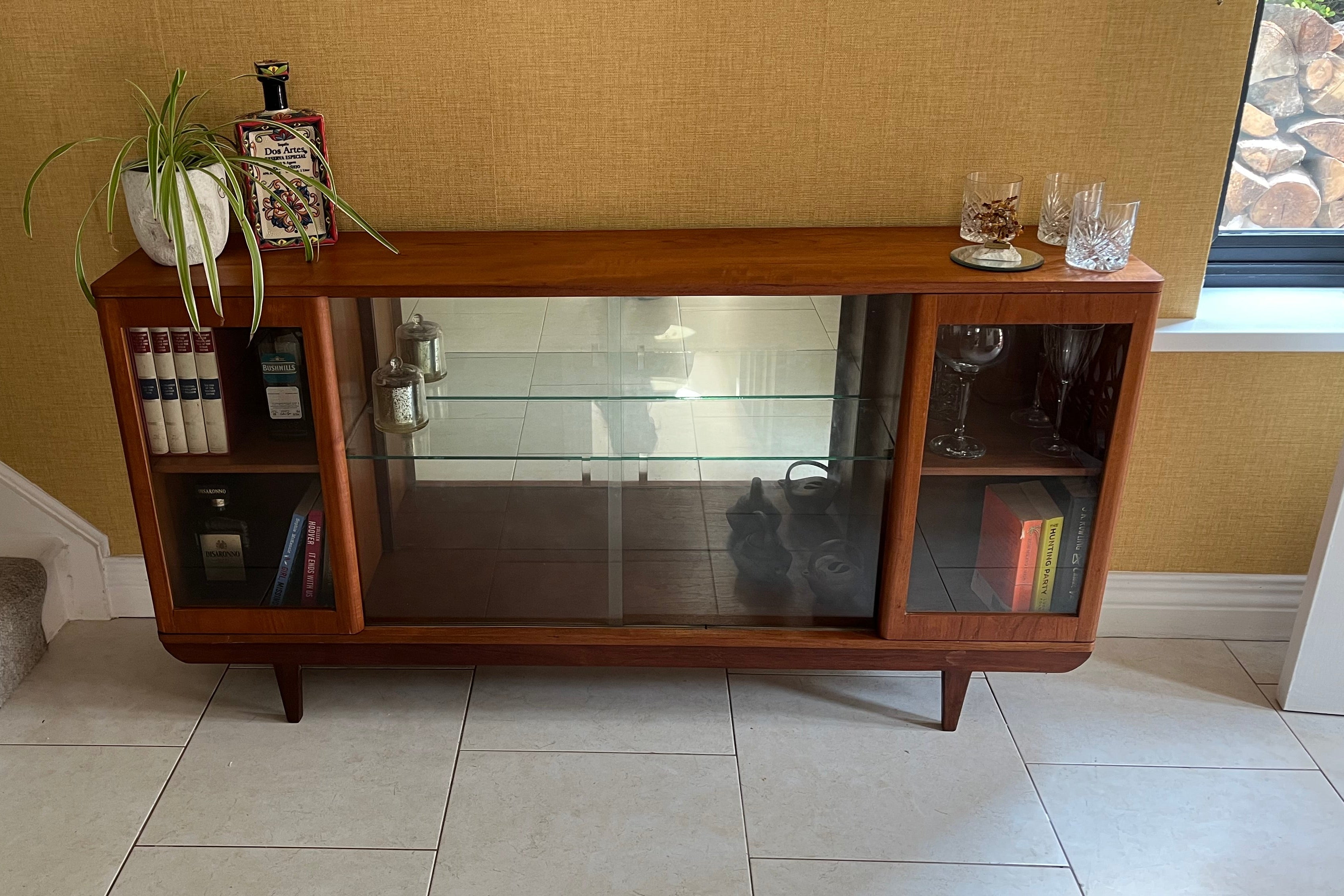 A mid-century modern SF Quinn wooden display cabinet with glass doors, shown in a home interior setting with tiled floors and textured walls by Grace & Joy Furniture