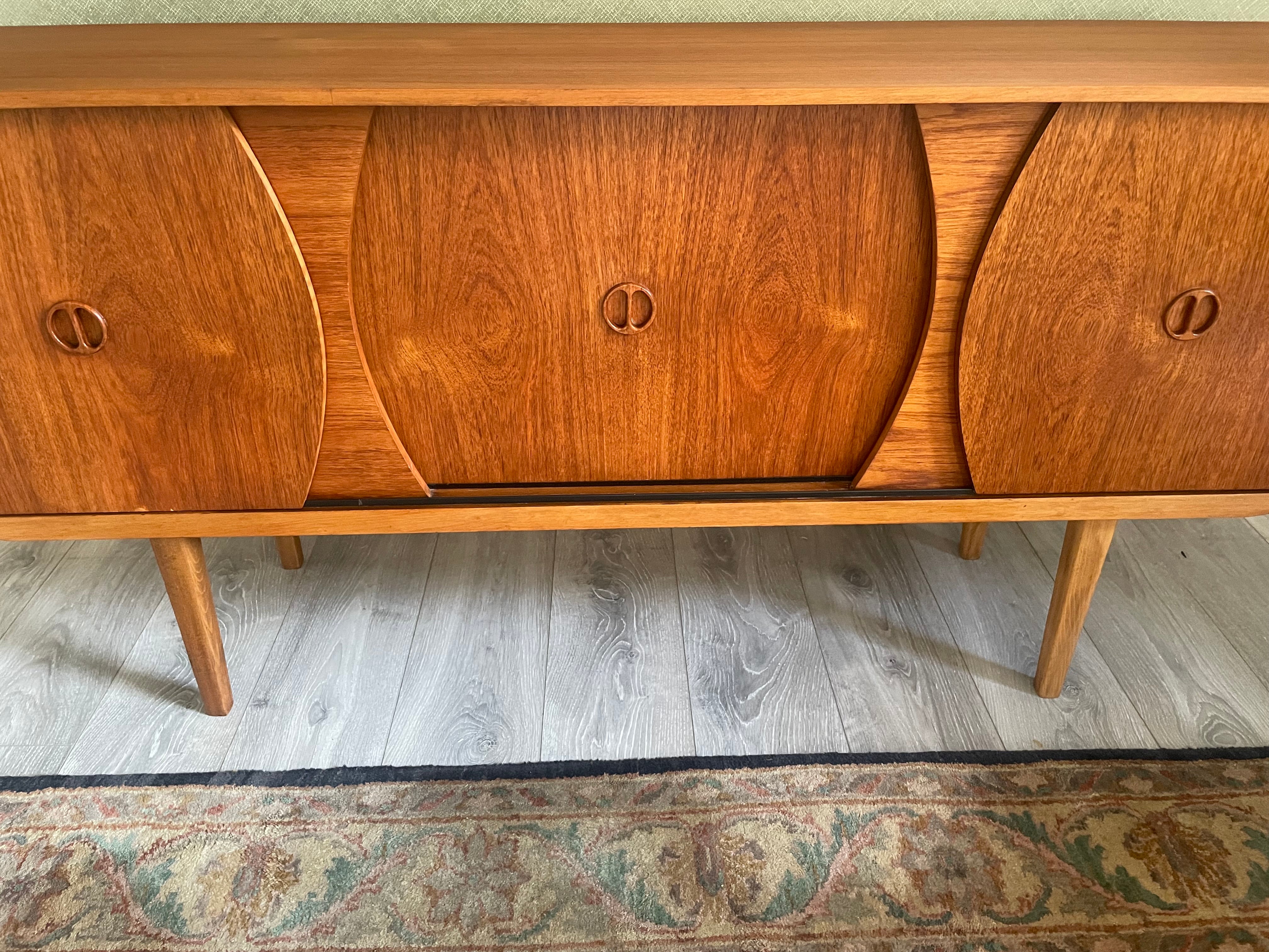 Restored mid-century modern wooden credenza or cabinet with three curved doors and circular pull handles, photographed in a home interior with wood flooring and a patterned rug visible by Grace & Joy Furniture