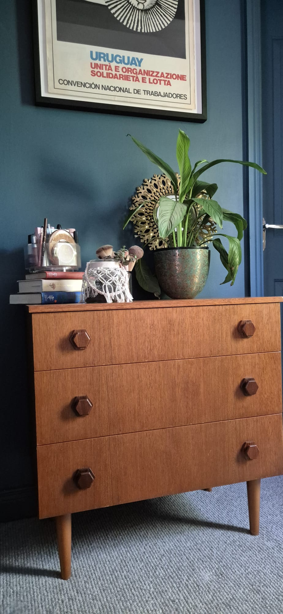 A mid-century modern wooden chest of drawers with three drawers and round dark knobs, styled with a potted plant, books, and a small decorative figure in a room with a dark blue wall by Grace & Joy Furniture