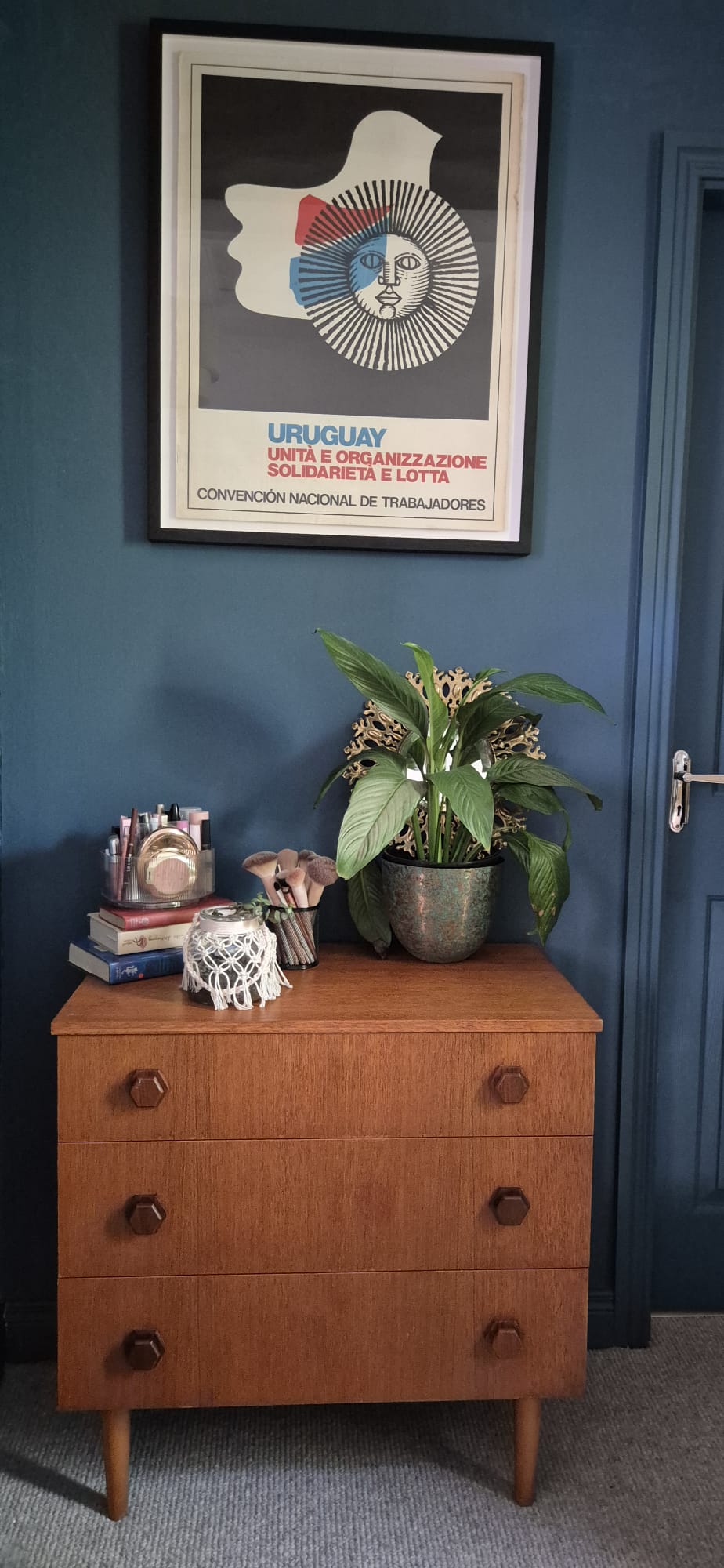 A mid-century modern wooden chest of drawers with three drawers and round dark knobs, styled with a potted plant, books, and a small decorative figure in a room with a dark blue wall by Grace & Joy Furniture